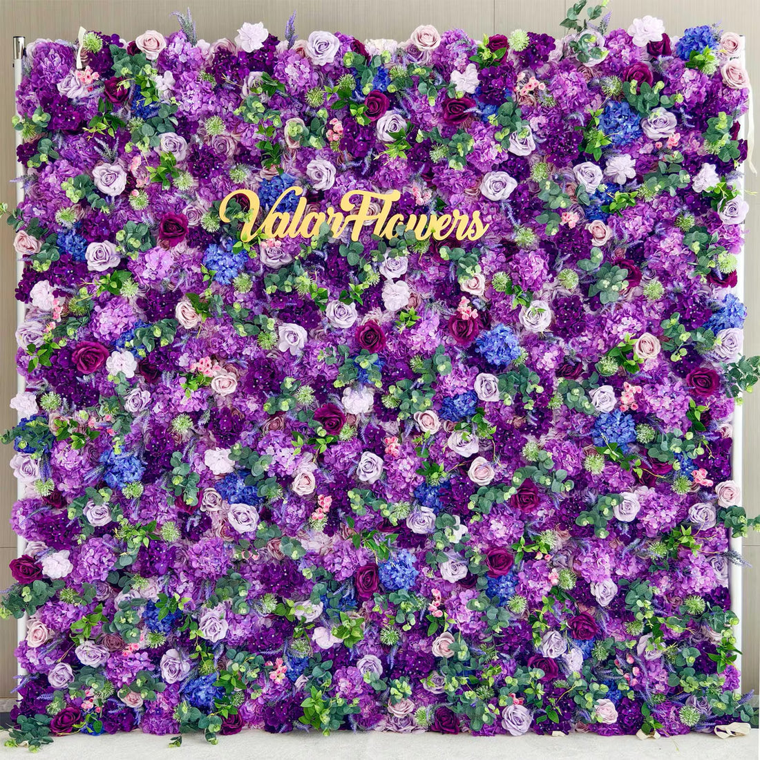 This image showcases a stunning floral wall by Valar Flowers, featuring a vibrant mix of purple, lavender, and blue blooms, along with some green foliage. The golden "Valar Flowers" logo stands out prominently. The dense arrangement of flowers creates a rich, luxurious, and visually captivating display, exuding elegance and natural beauty, perfect for decorative purposes in events or as a striking backdrop.
