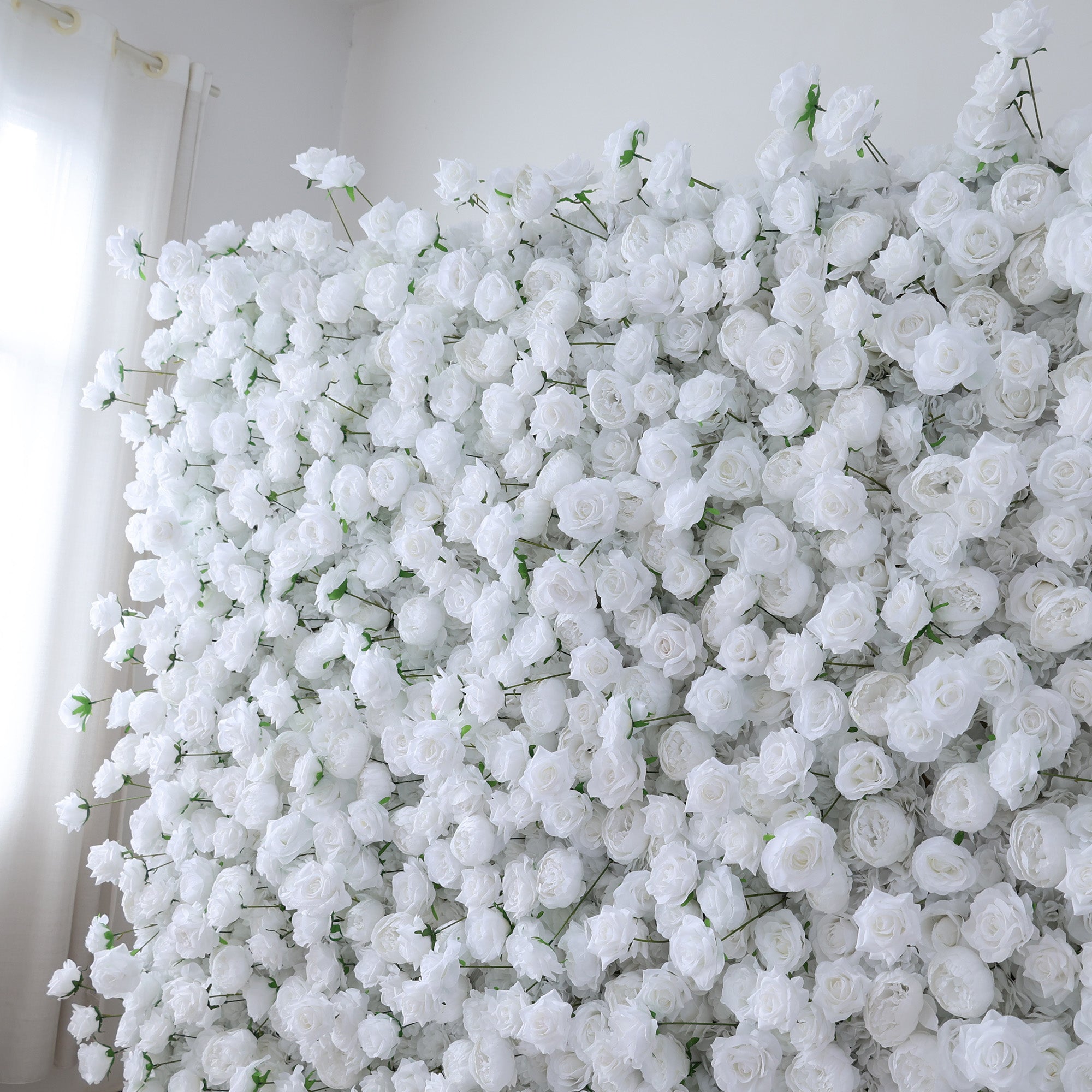 This image showcases a dense arrangement of white artificial roses, forming a lush and voluminous floral display. The flowers are tightly packed, with their soft, creamy petals and subtle green stems creating a sense of purity and elegance. Set indoors, likely near a window with light filtering through, the scene exudes a serene and sophisticated atmosphere. It&