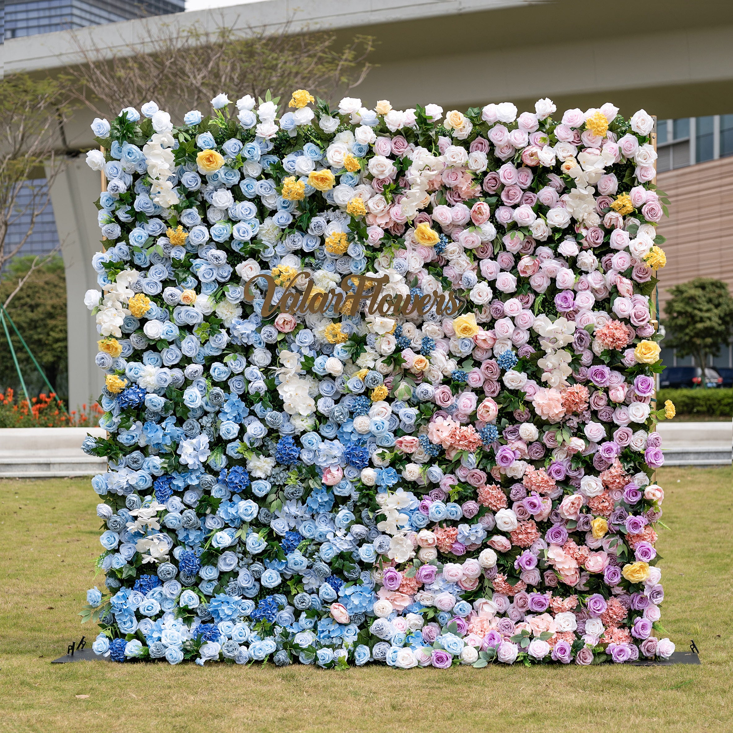 This is a stunning gradient artificial flower wall backdrop from Valar Flowers, set outdoors on a grassy lawn with an urban skyline in the background. It features a lush, dense arrangement of lifelike blooms: soft sky-blue delphiniums at the bottom, transitioning upward to creamy white roses, sunny yellow poppies, blush pink peonies, and lavender roses. The seamless color gradient creates an elegant, romantic focal point perfect for weddings, photo booths, and luxury event decor.