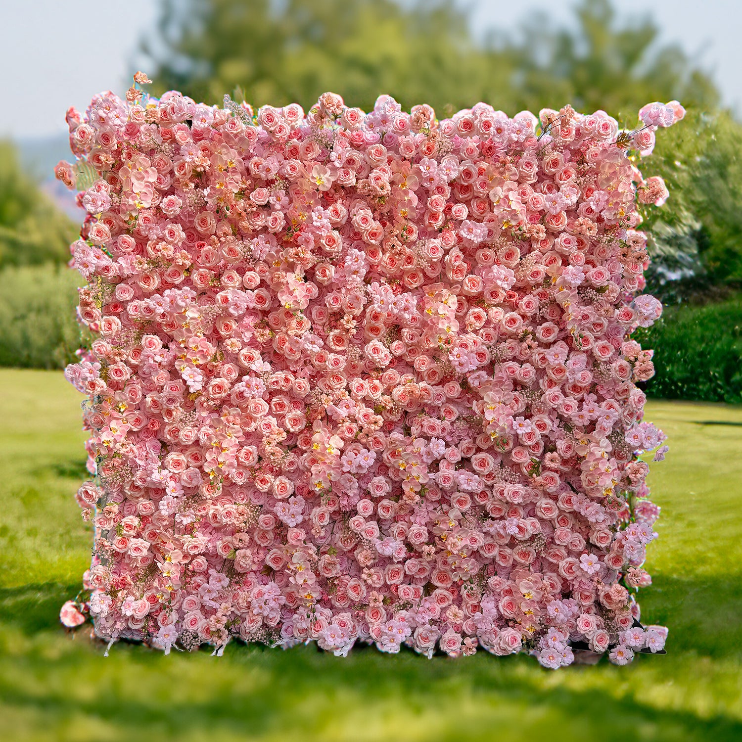 This photo features a large, square floral backdrop densely packed with soft pink roses, delicate pink orchids, and small pink flowers. Set on a grassy field with greenery in the background, it exudes a serene and romantic charm. Ideal for events like a "Pink Blossom Garden Party" or an intimate wedding, it creates a dreamy and picturesque atmosphere.