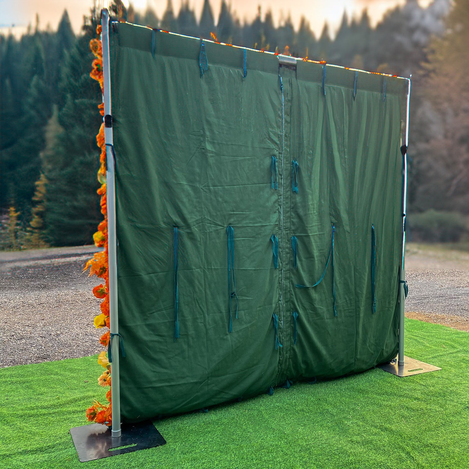 This image depicts a setup featuring a green fabric backdrop supported by a metal frame with square bases. The frame is adorned on one side with a vibrant arrangement of artificial flowers in shades of orange and yellow, likely roses and other assorted blooms. The style is functional with a touch of decorative flair, suitable for creating a partition or backdrop for events.