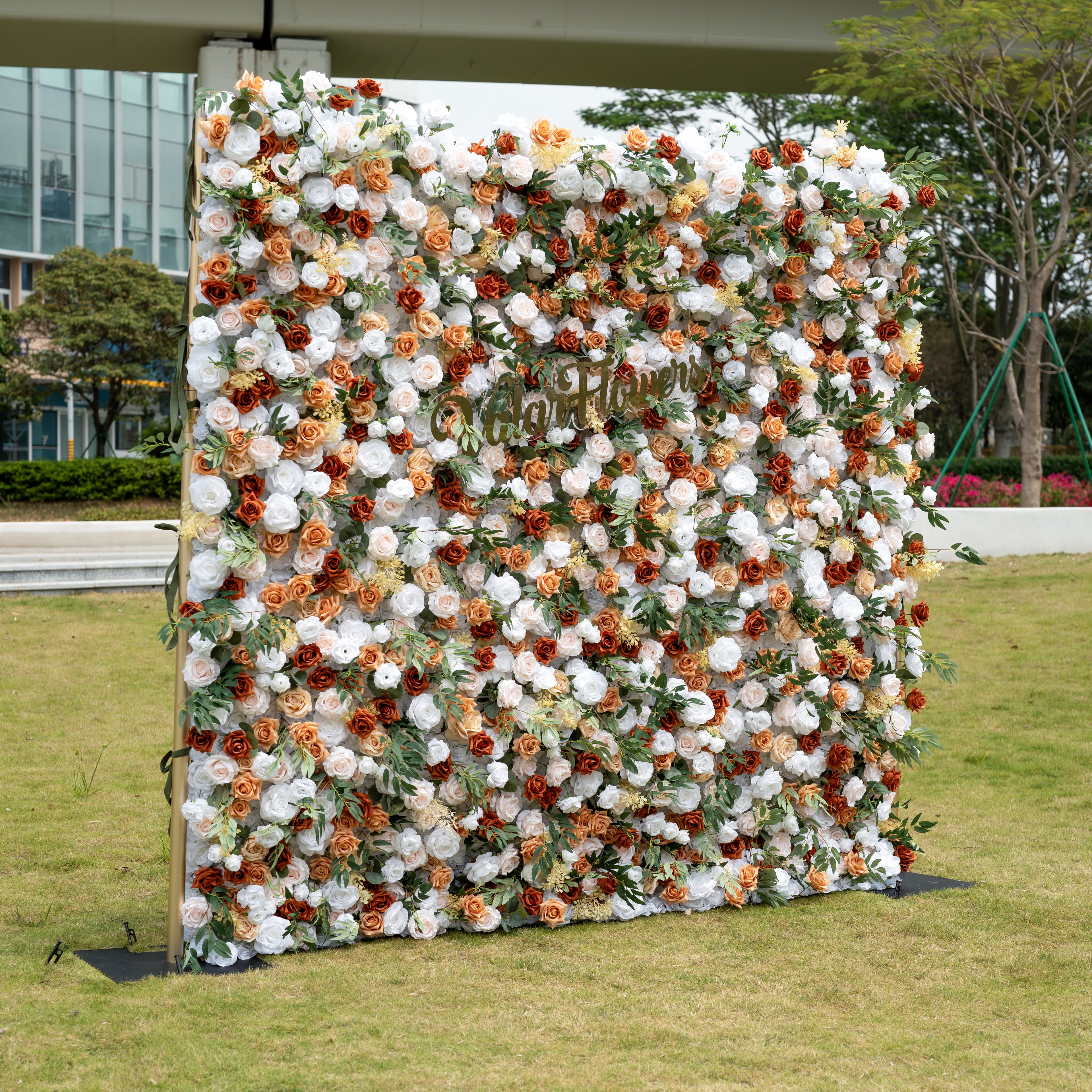 This is ValarFlowers’ premium terracotta & cream artificial flower wall backdrop, set outdoors on a grassy lawn with an urban skyline in the background. It features a lush, dense arrangement of lifelike blooms: burnt orange roses, creamy white peonies, blush pink roses, and vibrant green foliage. This warm, timeless, reusable floral display creates an elegant, romantic focal point ideal for rustic weddings, fall events, photo booths, and luxury outdoor decor.