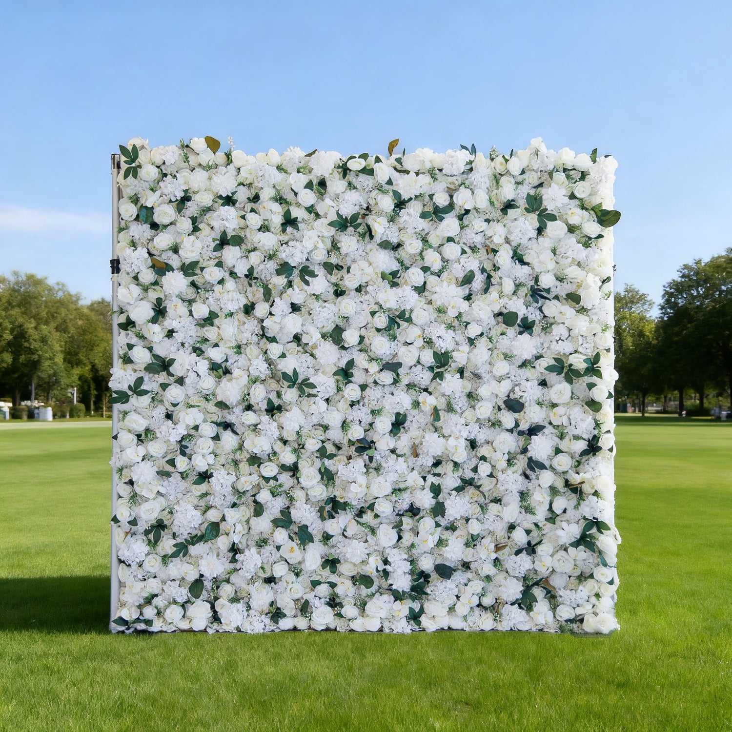 This image features a large, square floral wall densely covered with white flowers, interspersed with green leaves. Set outdoors on a lush green lawn under a clear blue sky, with trees in the background, it creates an elegant and serene atmosphere. Though "Valar Flowers" branding isn&