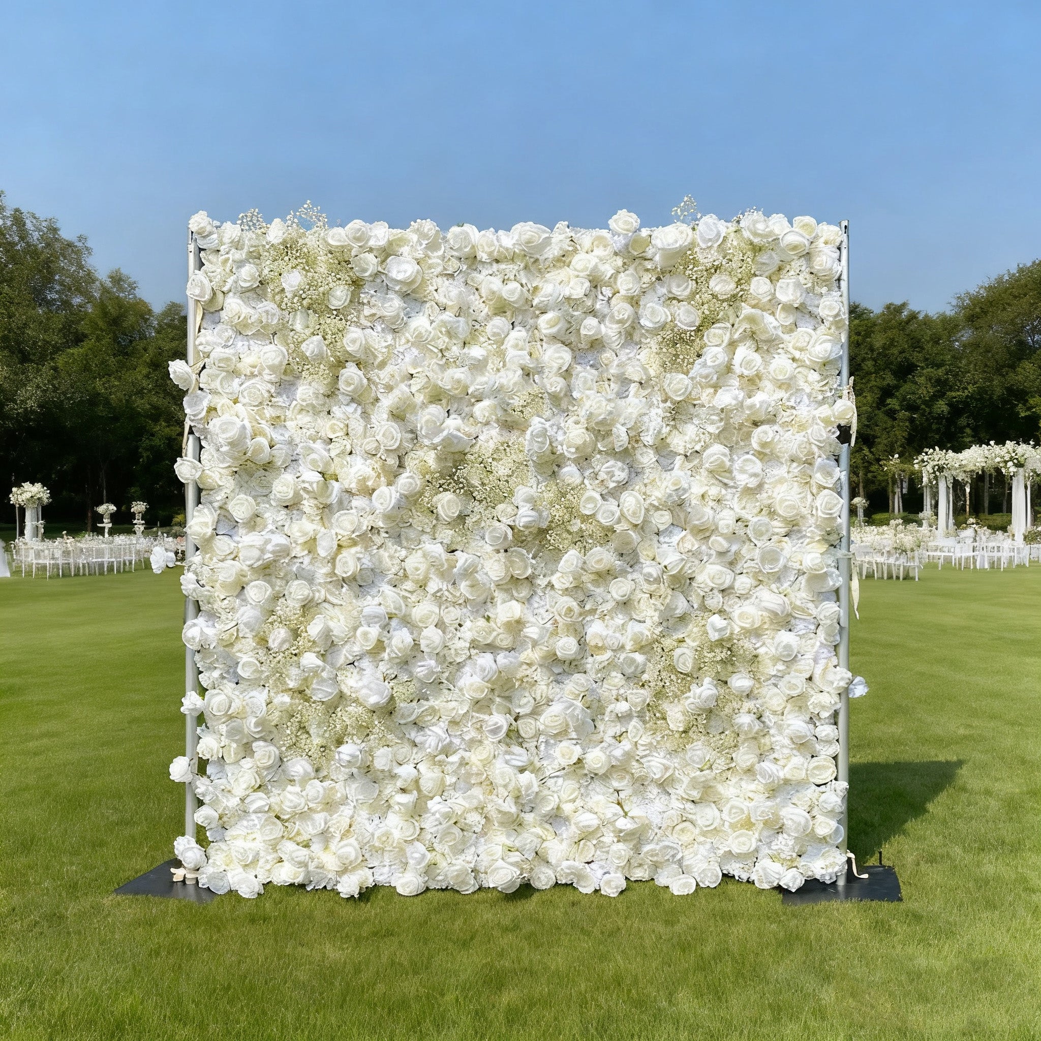 This image features a large, square floral wall composed of abundant white flowers. Set outdoors on a lush green lawn under a clear blue sky, with event setups like white chairs and decorative structures in the background, it creates an elegant and serene atmosphere, perfect for occasions such as weddings or other special events.