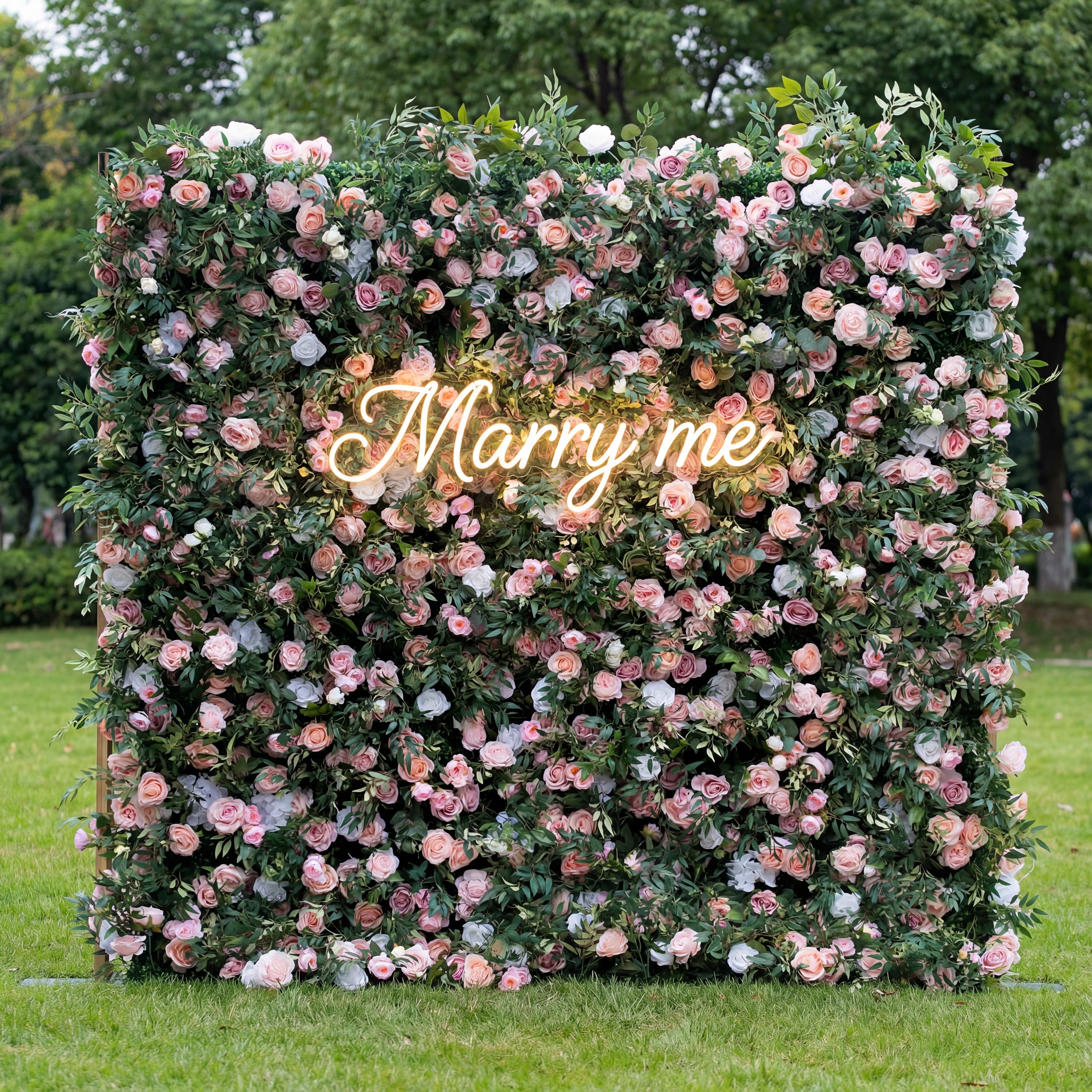 This romantic floral wall (set on sunlit grass, tree backdrop) blends soft pink, blush, and white blooms, interwoven with lush green foliage for texture. A glowing cursive "Marry me" neon sign (pale white) anchors the center, glowing gently against the muted, dreamy palette. Natural light enhances its airy, intimate vibe—ideal for proposals, merging heartfelt charm with elegant, lush softness as a warm, eye-catching focal point.