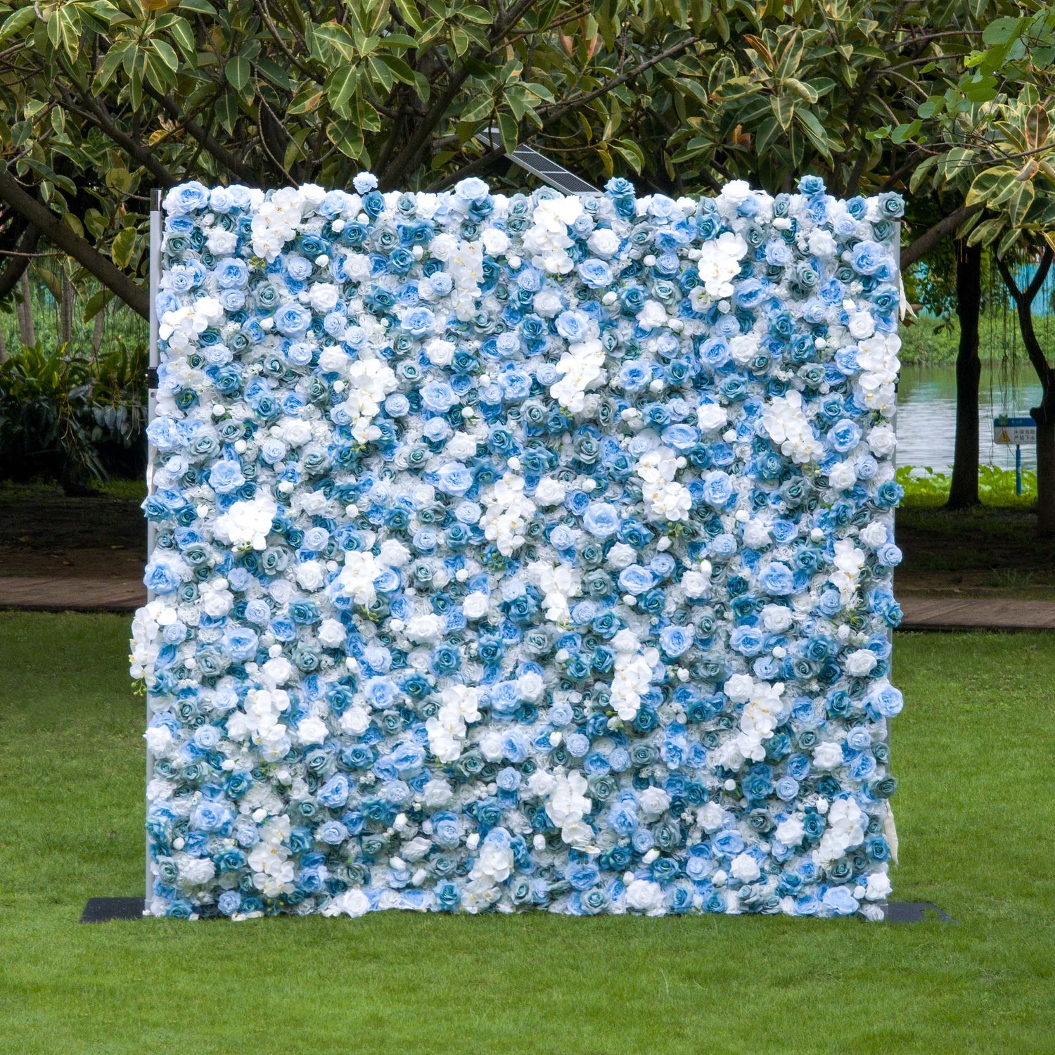 This photo showcases a large, eye - catching floral backdrop densely adorned with blue and white roses, along with white orchids. The lush arrangement creates a dreamy, textured appearance. Set outdoors on a grassy area with trees and a body of water in the background, it exudes a serene and elegant ambiance. Ideal for weddings, garden parties, or upscale outdoor events, it serves as a stunning decorative element.