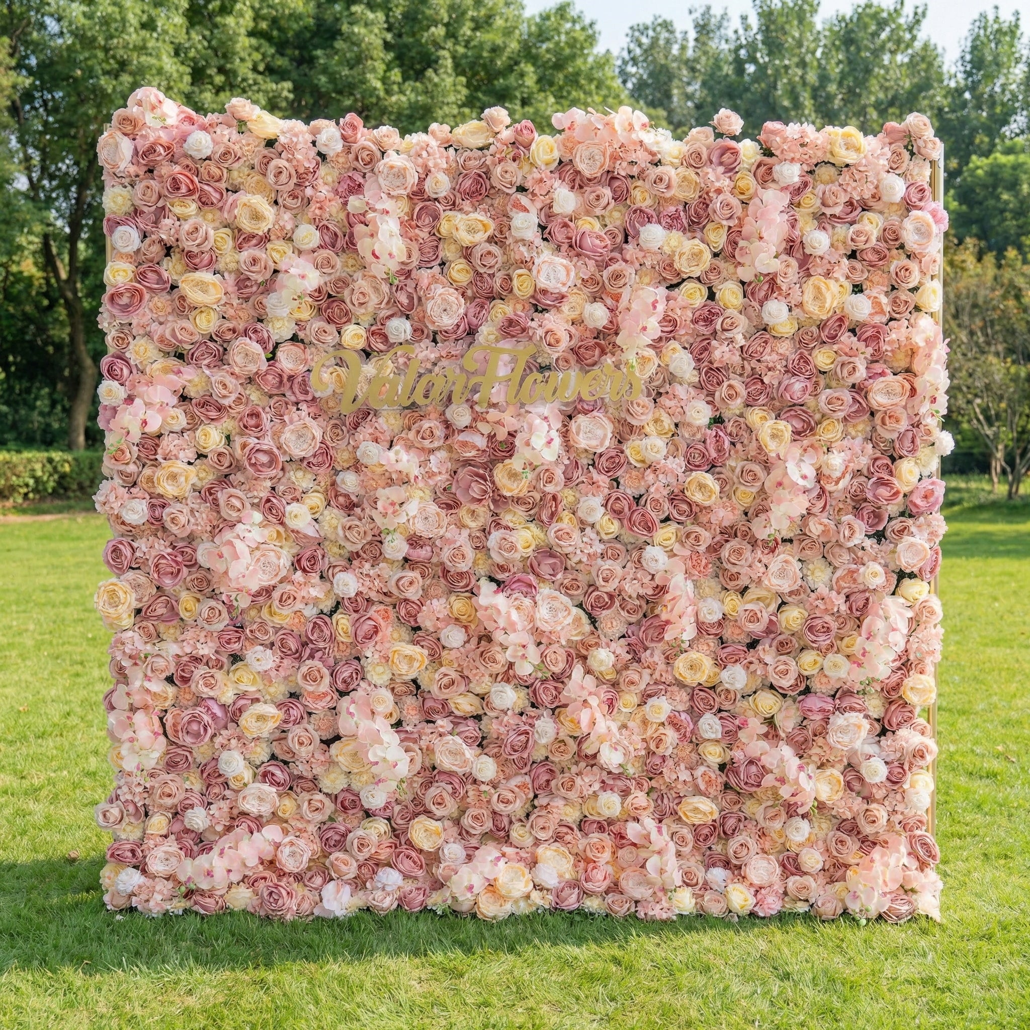 This stunning outdoor floral wall from Valar Flowers is a lush tapestry of blush pink, creamy white, and soft peach blooms—including roses, peonies, and delicate filler flowers. Set on a sunlit green lawn framed by leafy trees, the dense, layered design creates a rich, three-dimensional texture, evoking a warm, romantic, and naturally elegant atmosphere ideal for weddings, photo backdrops, or elegant outdoor celebrations.