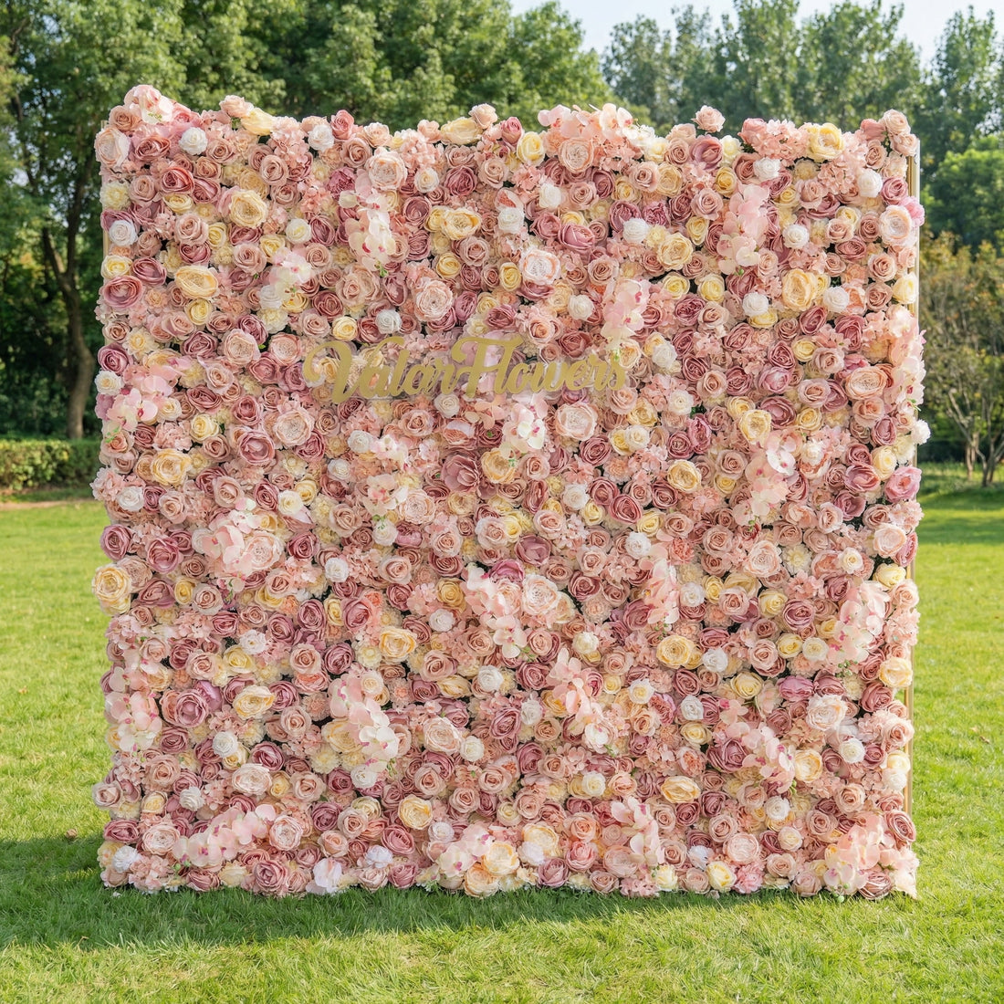 This stunning outdoor floral wall from Valar Flowers is a lush tapestry of blush pink, creamy white, and soft peach blooms—including roses, peonies, and delicate filler flowers. Set on a sunlit green lawn framed by leafy trees, the dense, layered design creates a rich, three-dimensional texture, evoking a warm, romantic, and naturally elegant atmosphere ideal for weddings, photo backdrops, or elegant outdoor celebrations.