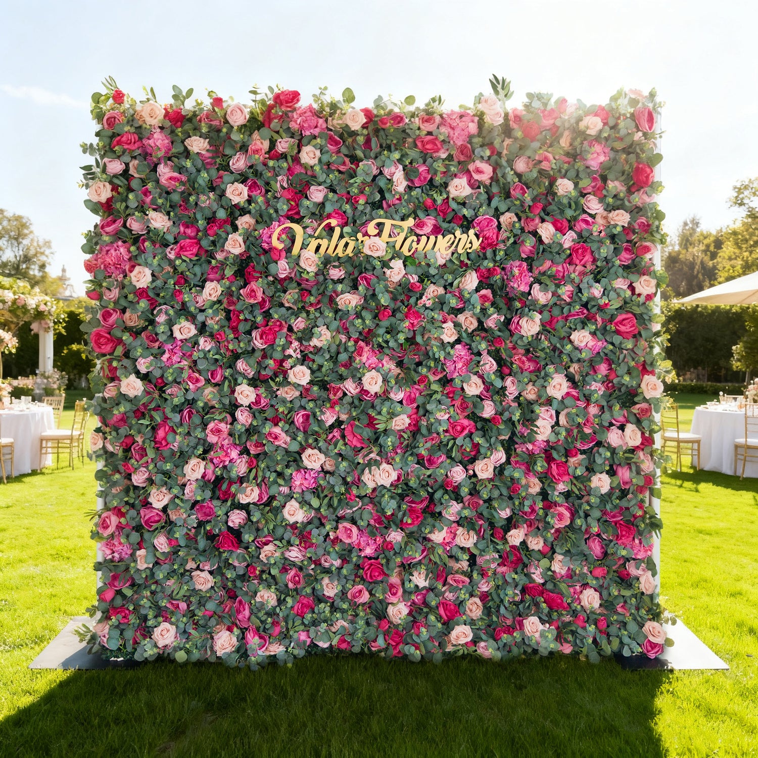 This image features a large, vibrant floral wall outdoors on a lush green lawn. Densely covered with roses in shades of pink, red, and white (interspersed with green foliage), it has a golden “Valar Flowers” sign at the center. The background includes event setups (tables, tents) under a bright, sunny sky with trees, exuding a cheerful, elegant vibe ideal for celebrations.