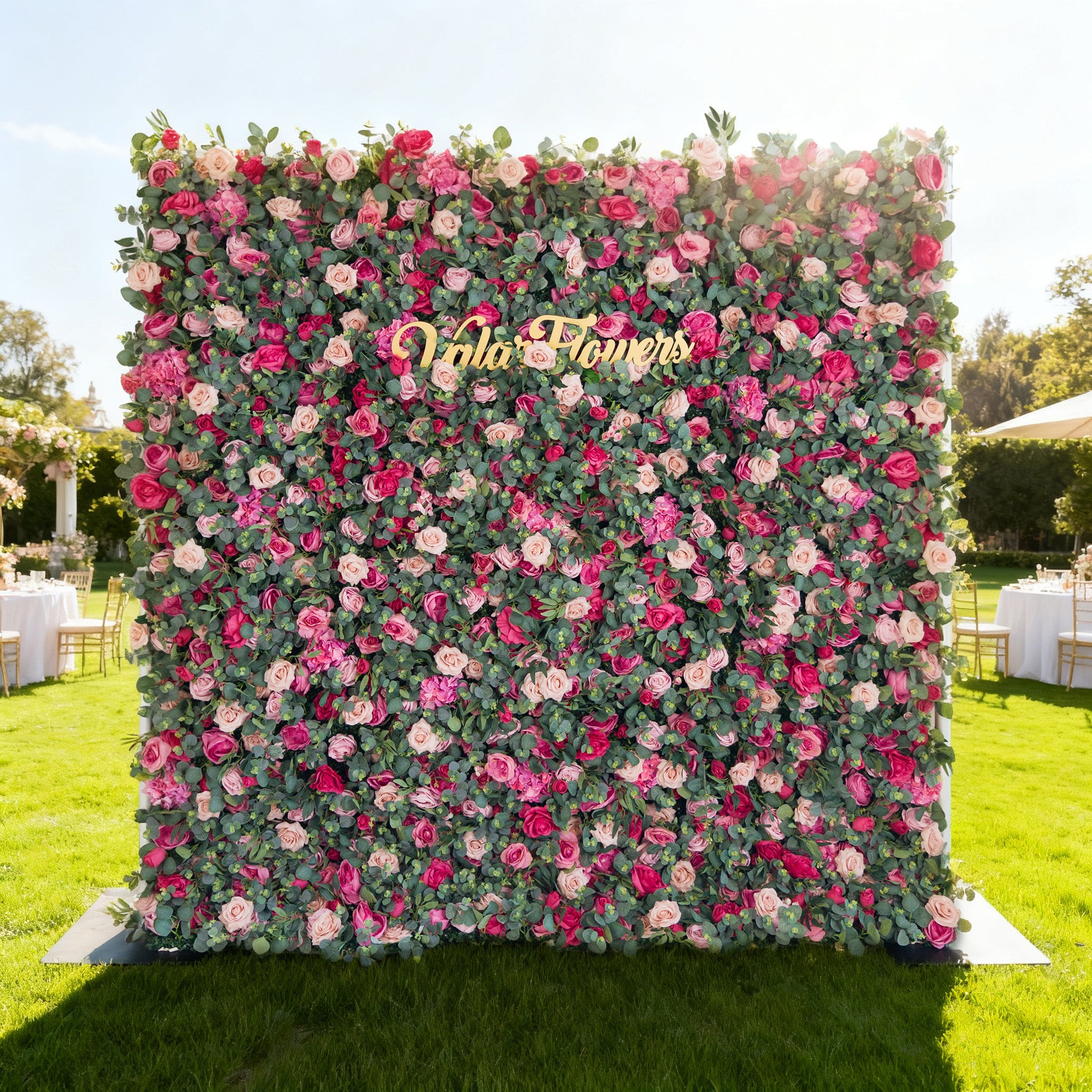 This image features a large, vibrant floral wall outdoors on a lush green lawn. Densely covered with roses in shades of pink, red, and white (interspersed with green foliage), it has a golden “Valar Flowers” sign at the center. The background includes event setups (tables, tents) under a bright, sunny sky with trees, exuding a cheerful, elegant vibe ideal for celebrations.