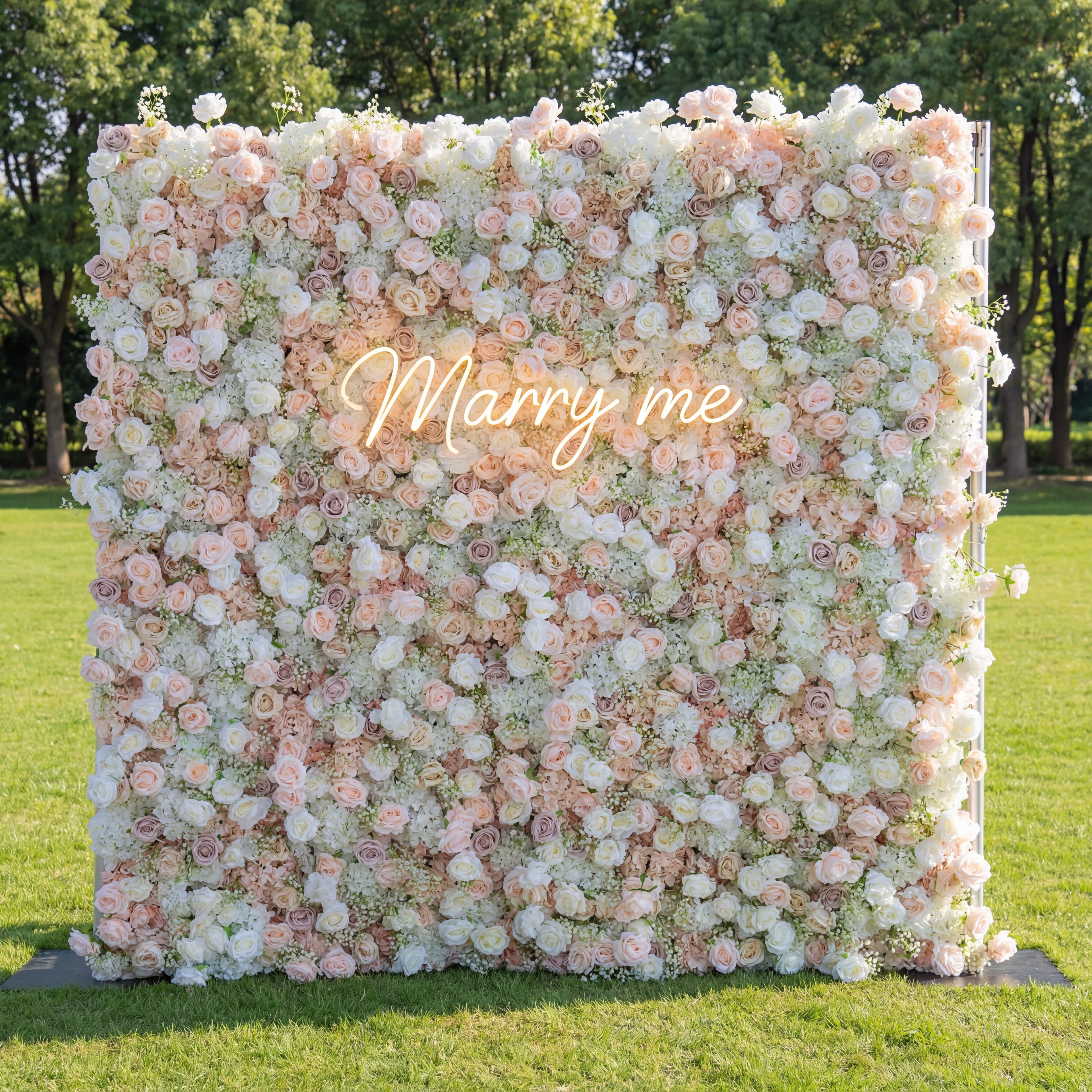 This outdoor backdrop (set on lush green grass, tree-framed) features a soft, dense wall of blush, ivory, and pale pink blooms—roses, peonies, and baby’s breath—creating an airy, romantic texture. A glowing "Marry Me" neon sign (cursive, light-lit) anchors the center, its gentle glow contrasting with the muted, elegant palette. The delicate display exudes warmth, perfect for outdoor proposals, garden weddings, or intimate romantic events.