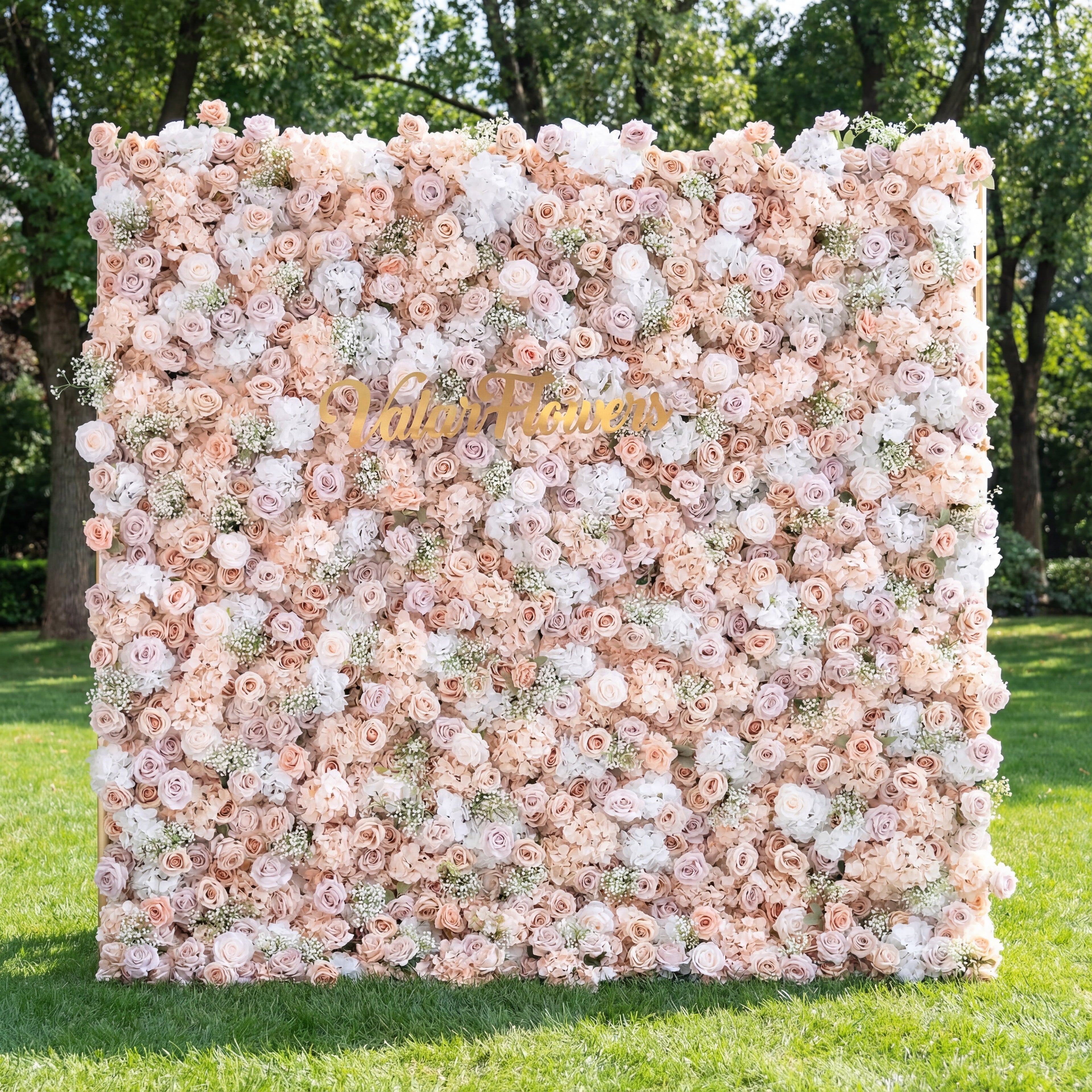 This outdoor backdrop (set on lush green grass, tree-framed) is a soft, dense wall of blush, ivory, and pale pink blooms—roses, peonies, and baby’s breath—creating an airy, romantic texture. A polished, cursive "Valar Flowers" gold sign anchors the center, contrasting gently with the muted, elegant palette. The delicate display exudes sophistication, perfect for wedding decor, garden events, or upscale photo setups.