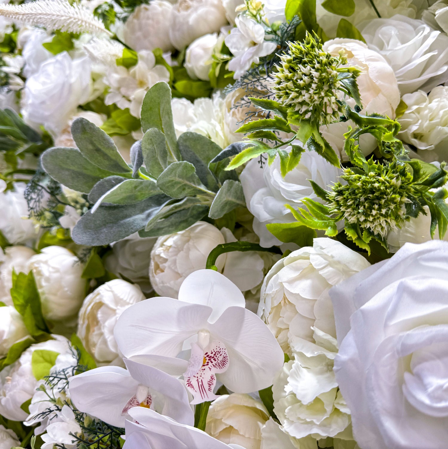 This close - up image showcases a variety of white and off - white flowers on a flower wall. The flowers are accompanied by green leaves, creating a lush and vibrant display. The close - up view highlights the intricate details of the petals, including the soft folds and natural - looking arrangements. The different types of flowers, such as roses and possibly peonies, add to the richness of the display.