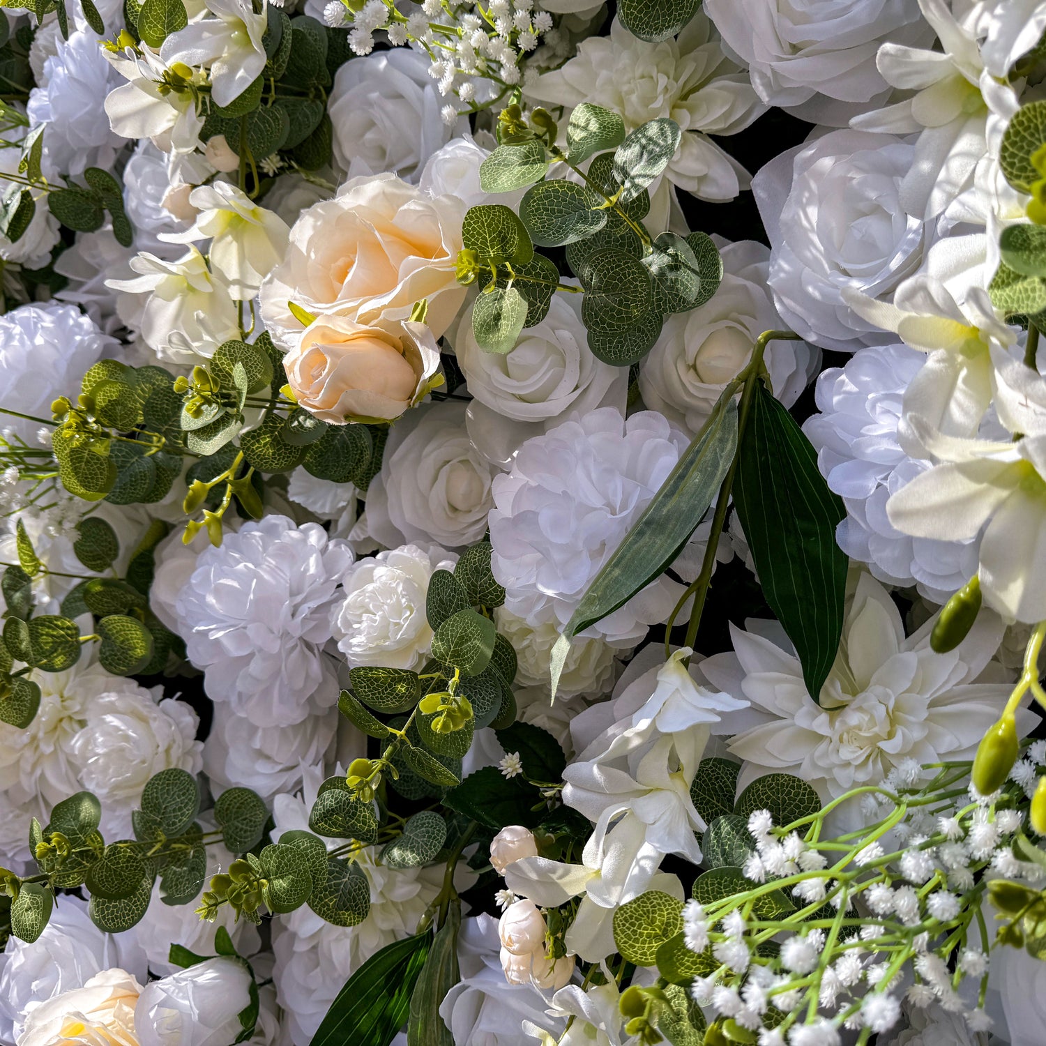 This artificial flower wall is a stunning piece. It is densely packed with a variety of white and off - white flowers, including roses and lilies. The flowers are complemented by lush green leaves and small floral buds, creating a rich and textured appearance. It&