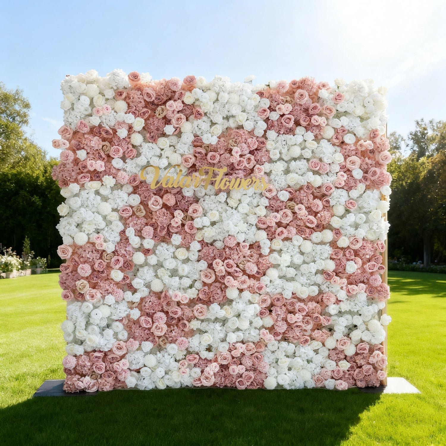 This image showcases a large floral wall with a lovely mix of pink and white flowers. "Valar Flowers" is displayed in golden text. Set outdoors on a lush green lawn under a light sky, with trees in the background, it creates a soft, elegant, and charming atmosphere, perfect for events or decorative displays.