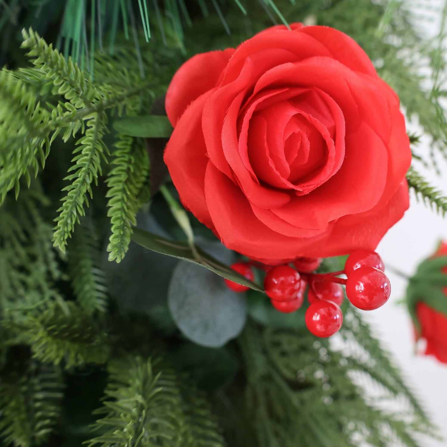 This close - up image features a vibrant artificial red rose with layered petals, exuding a lifelike charm. Accompanying the rose are glossy red berries, adding a touch of brightness. They are nestled among lush green foliage, including fern - like leaves and other leafy textures. The combination creates a visually appealing and festive arrangement, ideal for decorative purposes, perhaps evoking a warm, celebratory atmosphere.
