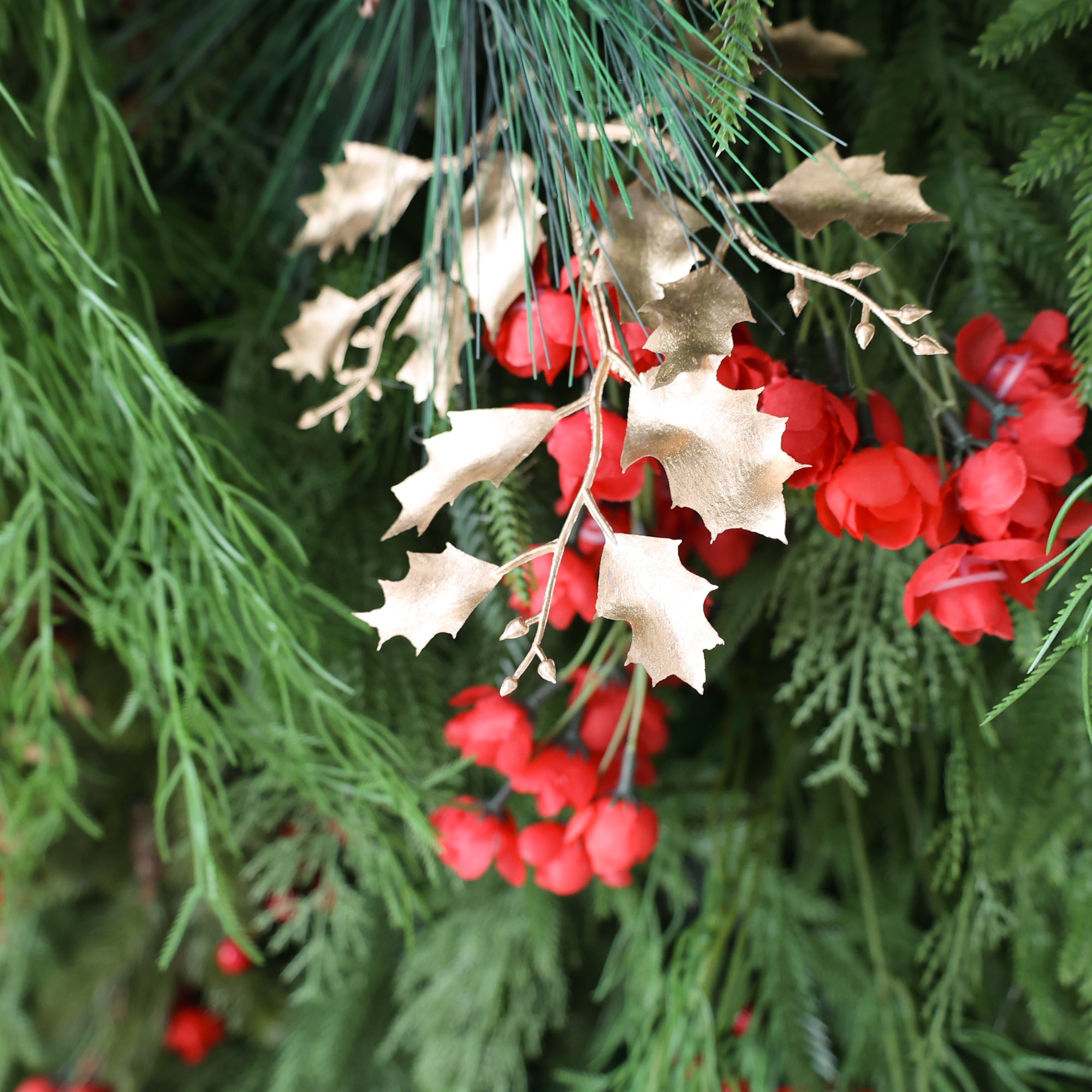 This close - up shows a festive arrangement with golden holly - shaped leaves and vibrant red berries. They are nestled among lush green foliage, including pine needles and fern - like leaves. The combination of gold, red, and green creates a warm, holiday - ready look, perfect for seasonal decorations.
