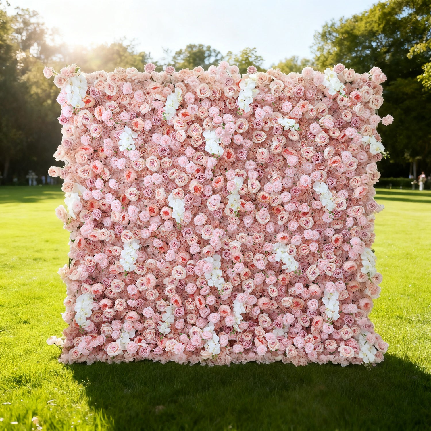This image showcases a beautiful floral wall by Valar Flowers, set outdoors on a lush green lawn. It features a dense arrangement of artificial pink and white flowers, creating a soft and romantic display. The backdrop is perfect for events, with the delicate hues and lush texture adding a touch of elegance, complemented by the sunny outdoor setting and surrounding greenery.