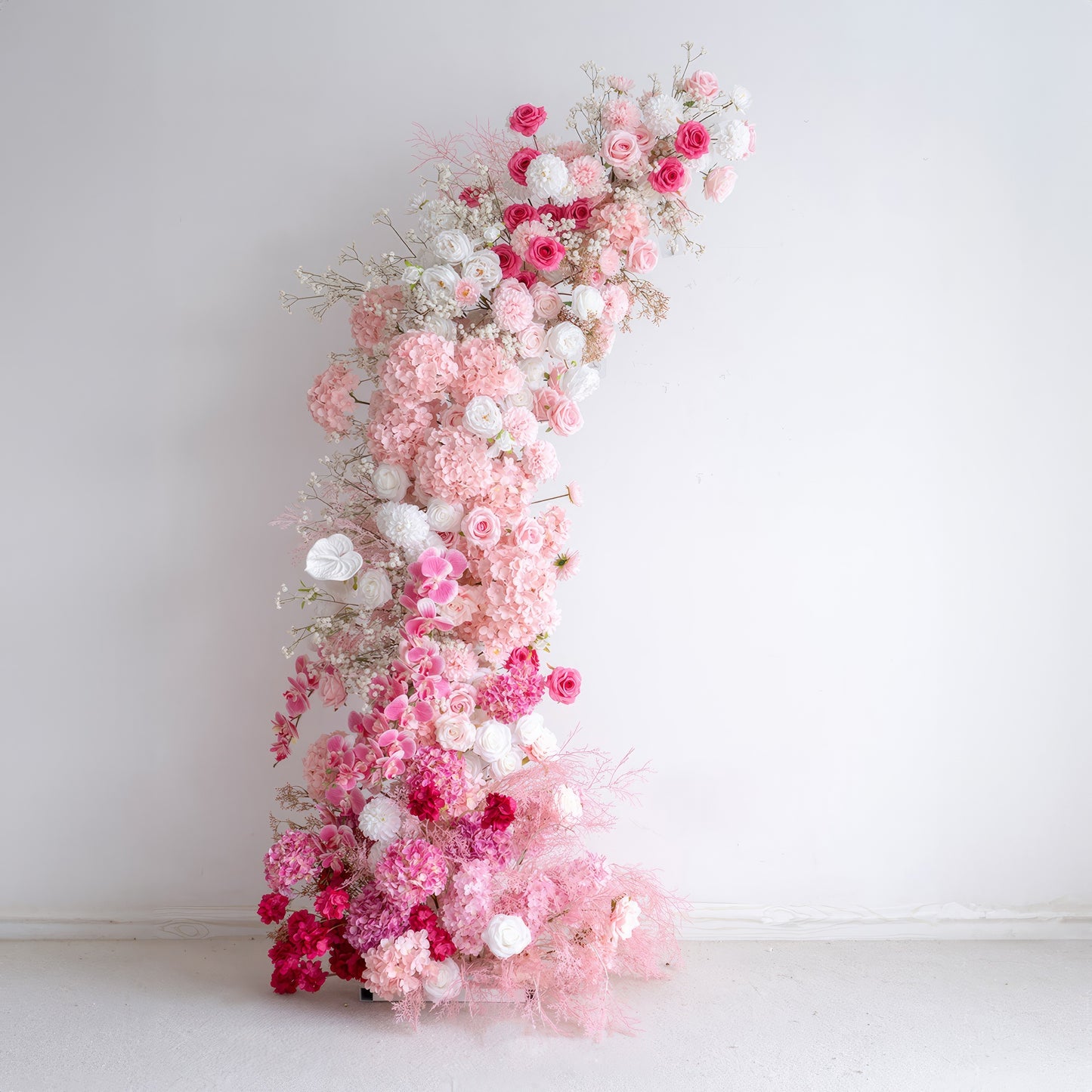This elegant, cascading floral arrangement features a romantic gradient of soft pink, deep rose, and white blooms, including hydrangeas, roses, orchids, and delicate filler flowers like baby's breath. The lush, layered design creates a dreamy, three-dimensional texture, with airy pink foliage adding a whimsical touch. Set against a minimalist white backdrop, it exudes timeless romance, making it a stunning focal point for weddings, engagement photoshoots, or romantic celebrations.