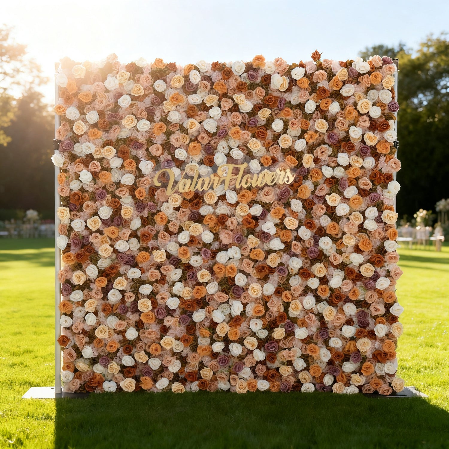 This image features a large, square floral wall composed of roses in warm tones like peach, cream, and light purple. In the center, golden text reads "Valar Flowers". Set outdoors on a lush green lawn, with sunlight casting a warm glow, it creates an elegant and inviting atmosphere, perfect for events or decorative displays.