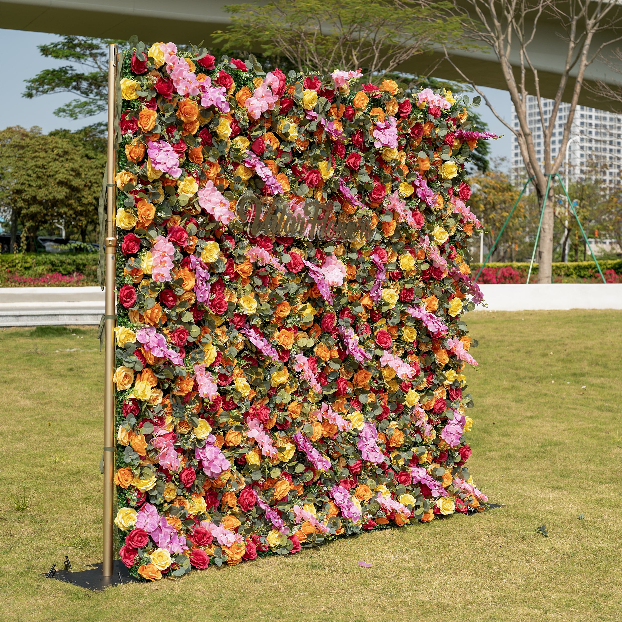 This is a full-size 8x8ft ValarFlowers premium multicolor artificial flower wall, mounted on an elegant gold adjustable backdrop stand. It features a lush, dense arrangement of lifelike burgundy, orange, yellow roses, pink hydrangeas, and realistic eucalyptus greenery, centered with a custom "ValarFlowers" logo sign. This high-quality, reusable floral display creates a vibrant, romantic focal point, ideal for wedding backdrops, photo booths, garden parties, and luxury outdoor event styling.