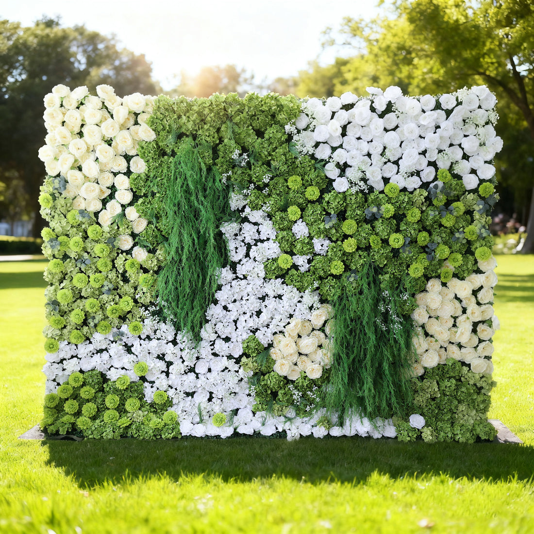 This image features a large, square floral wall outdoors on a lush green lawn. It combines various white flowers with abundant green foliage and trailing vines, creating a vibrant, natural, and refreshing display. Bathed in sunlight, with trees in the background, the scene exudes a sense of vitality, harmony, and natural beauty, perfect for decorative purposes in events or as a picturesque backdrop.