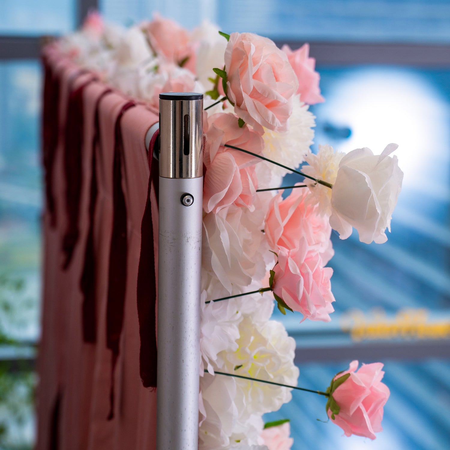 This image shows a close - up of a floral arrangement. There are pink and white roses with some green leaves, attached to a structure. The background includes a blurred view of a building through a window.