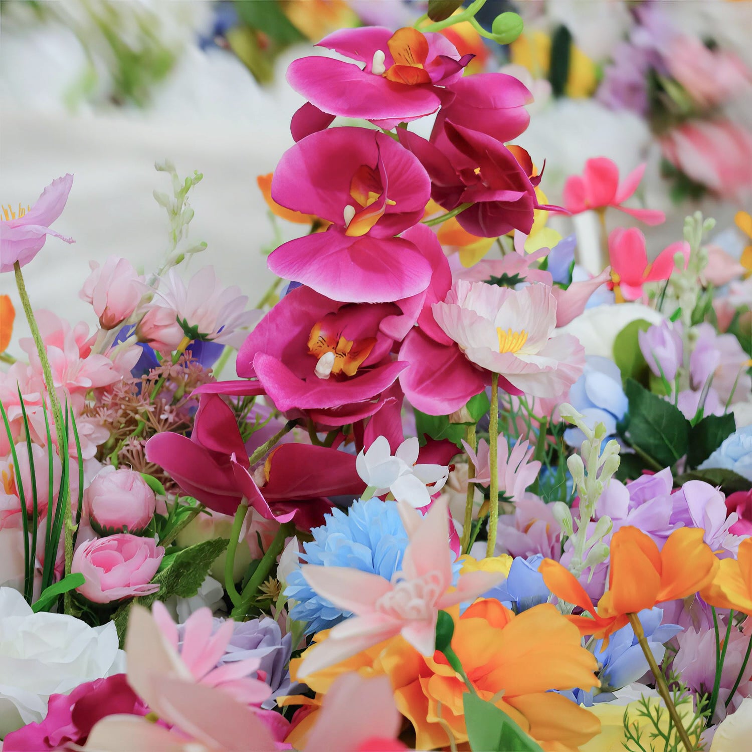 This image showcases a vibrant and colorful arrangement of artificial flowers. The palette includes vivid shades of pink, orange, blue, and white. Prominent flower types are orchids in deep magenta, along with delicate pink lilies, blue carnation - like blooms, and bright orange poppy - like flowers. The overall style is lively and cheerful, creating a lush and eye - catching display.