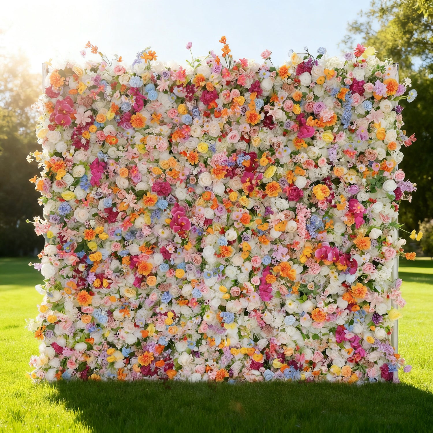 This image showcases a large, vibrant floral wall brimming with an array of colorful flowers in shades of pink, orange, yellow, blue, and white. Set outdoors on a lush green lawn under bright sunlight, it creates a lively and cheerful atmosphere, perfect for decorative displays or events, exuding a sense of natural beauty and joy.