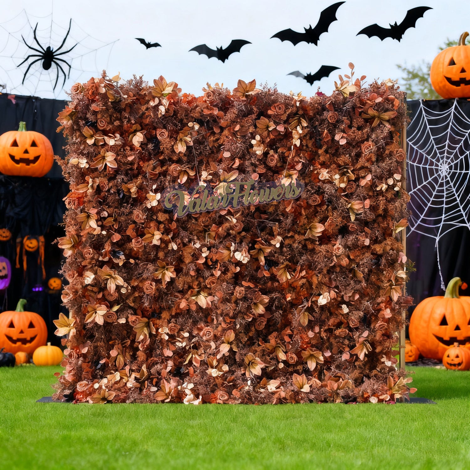 This image showcases a Halloween - themed floral wall with a rich, autumnal brown hue. "Valar Flowers" is displayed in golden text. Surrounding it are classic Halloween decorations like jack - o&