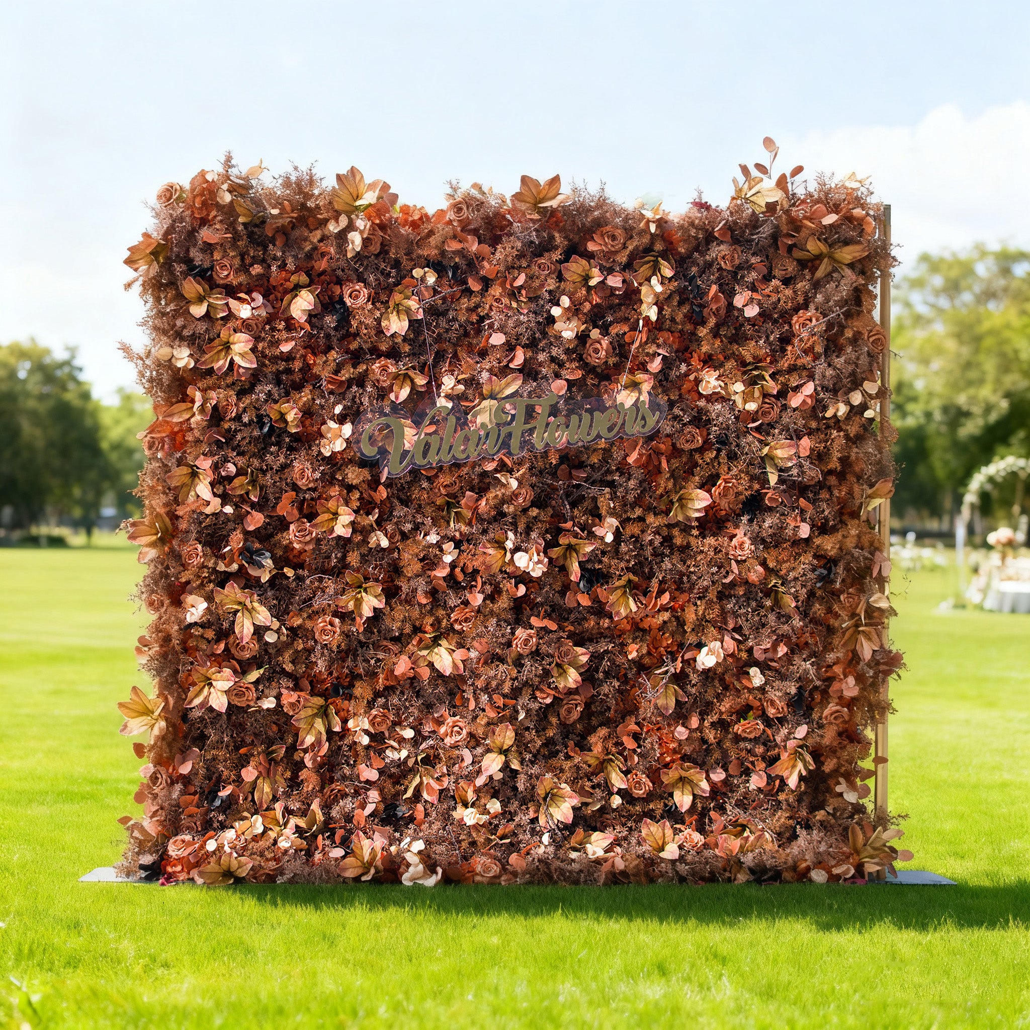 This image showcases a stunning floral backdrop by ValarFlowers, set outdoors on a vibrant lawn. It features a dense arrangement of artificial flowers and foliage in rich, warm tones of copper, bronze, and terracotta, creating a luxurious, textured display. The prominent “ValarFlowers” logo adds a branded touch. Bathed in sunlight with a scenic park and event setup in the background, this piece exudes elegance and sophistication, perfect for adding a lavish, nature-inspired element to outdoor events.