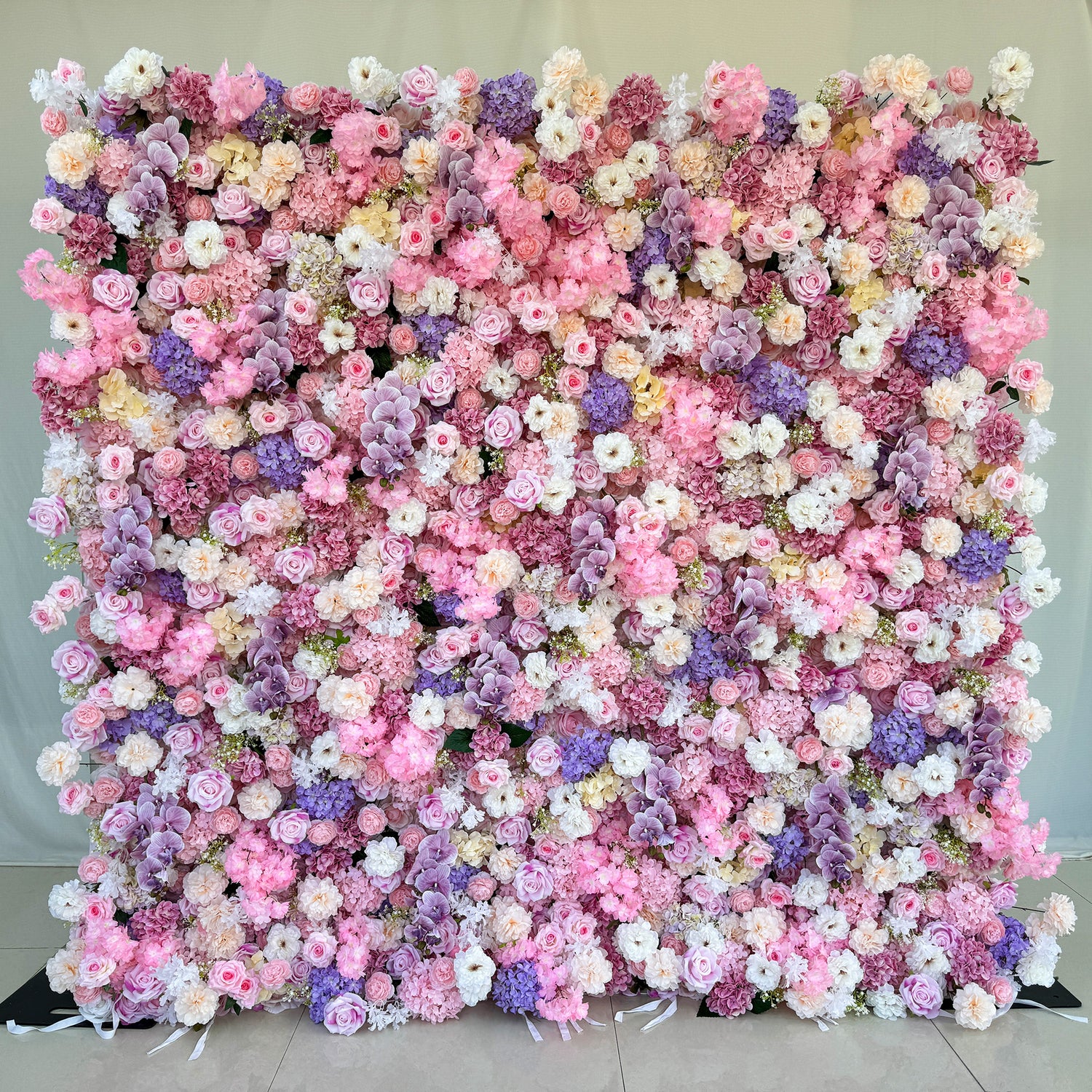 This photo from ValarFlowers showcases a large, dreamy floral backdrop. It is densely covered with an assortment of roses in soft pink, white, and hints of purple. Hydrangeas in shades of purple and pink add fullness, along with delicate carnations in white and light pink. Small white flowers and green foliage complete the arrangement. Ideal for weddings, bridal showers, or romantic events, it exudes elegance and charm.
