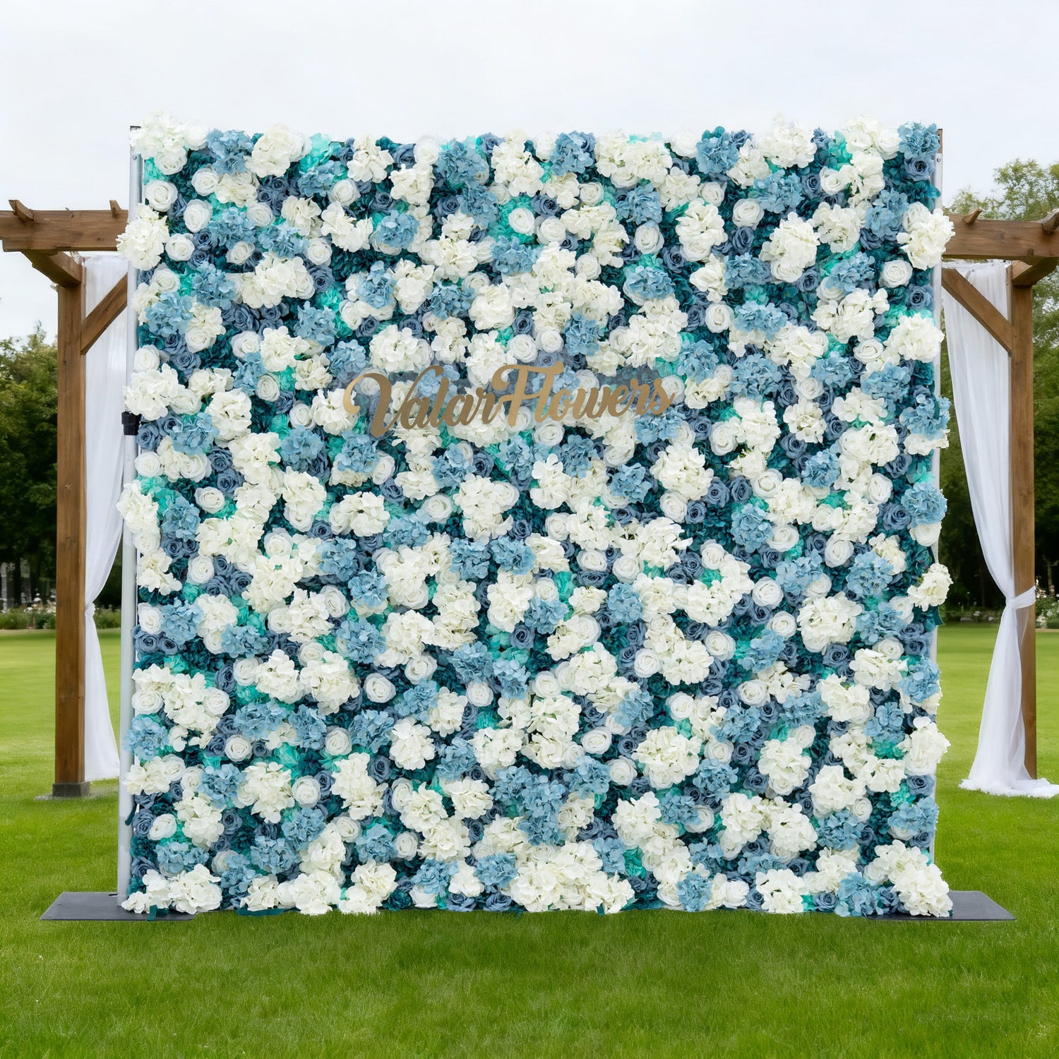 This image features a large floral wall composed of white and blue flowers. "Valar Flowers" is displayed in golden text on it. Set outdoors on a lush green lawn, the scene includes a wooden arch with white drapes, creating an elegant and serene atmosphere, perfect for events or decorative displays.