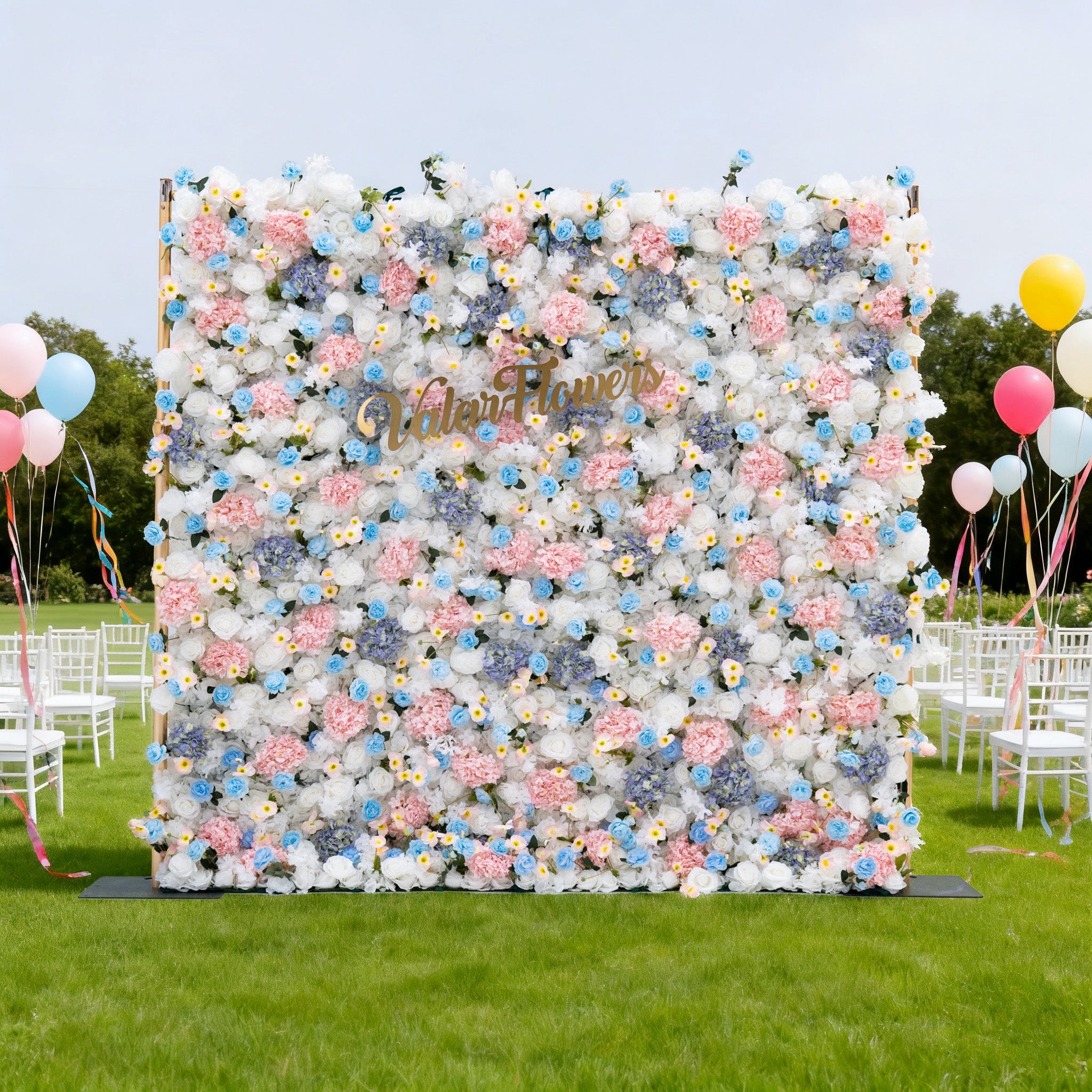 This image features a large floral wall with a soft palette of white, pink, and blue flowers. "ValarFlowers" is displayed in golden text on it. Set outdoors on a lush green lawn, the scene includes white chairs and colorful balloons, creating a cheerful and elegant atmosphere, perfect for events like weddings or celebrations.