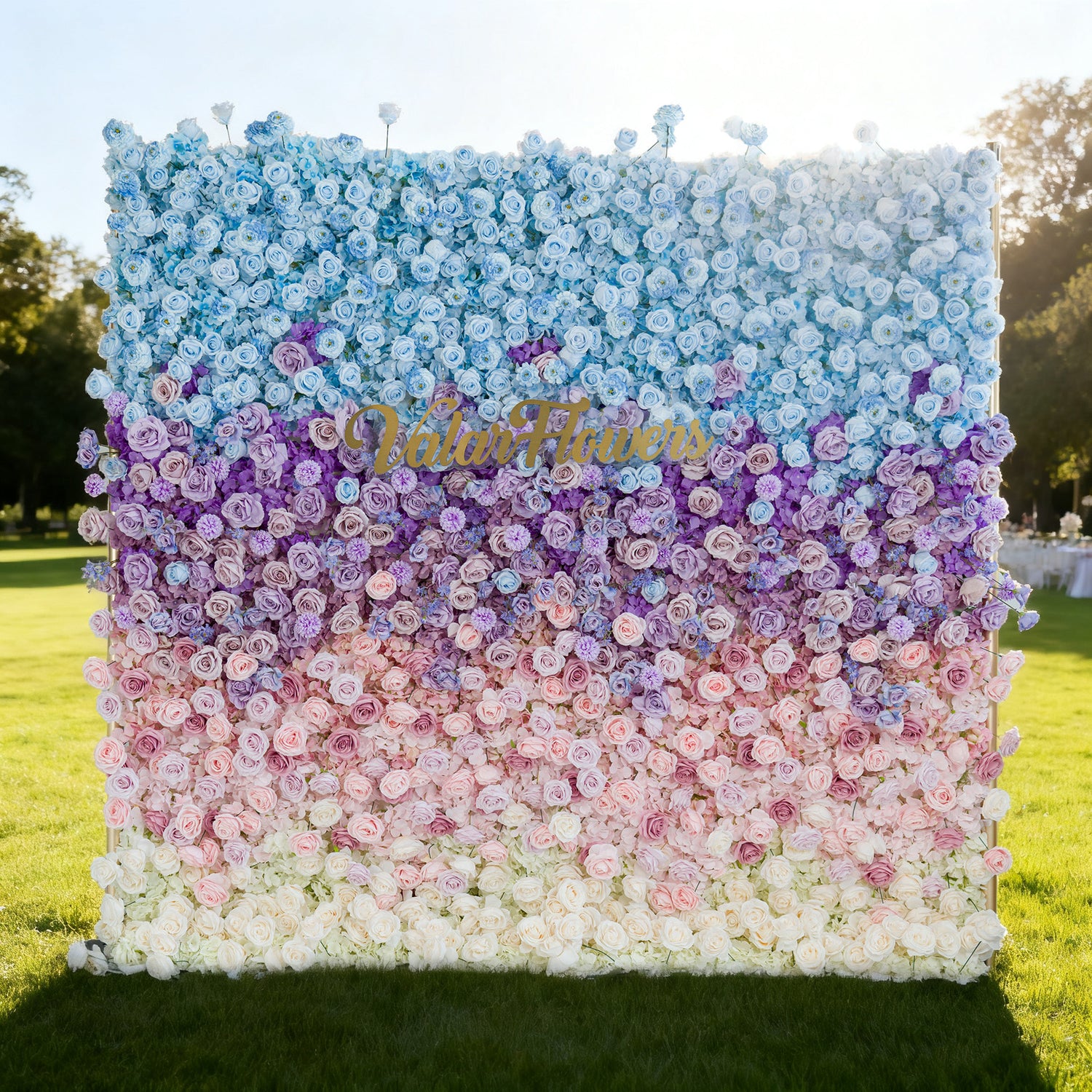 This image features a large floral wall with a gradient of colors from blue at the top, transitioning through purple, pink, to white at the bottom. "Valar Flowers" is displayed in golden text. Set outdoors on a lush green lawn under a bright sky, it creates a vibrant and elegant atmosphere, perfect for events or decorative displays.