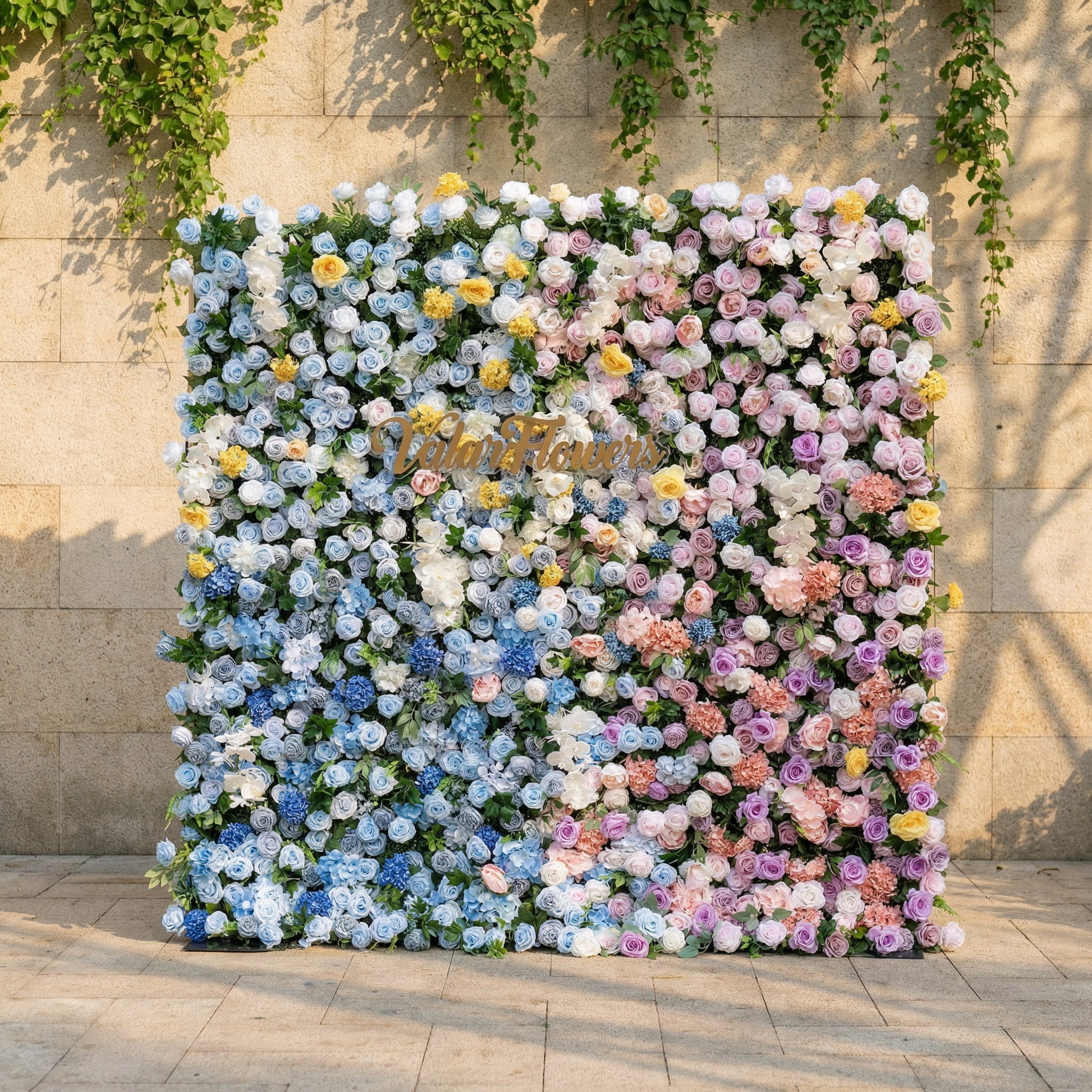 This is a stunning, gradient-style artificial flower wall backdrop from Valar Flowers, set against a sunlit stone wall with trailing ivy. It features a lush, dense arrangement of lifelike blooms: soft sky-blue delphiniums at the bottom, transitioning upward to creamy white roses, blush pink peonies, sunny yellow poppies, and lavender roses. The seamless color gradient creates an elegant, romantic focal point perfect for weddings, photo booths, and luxury event decor.