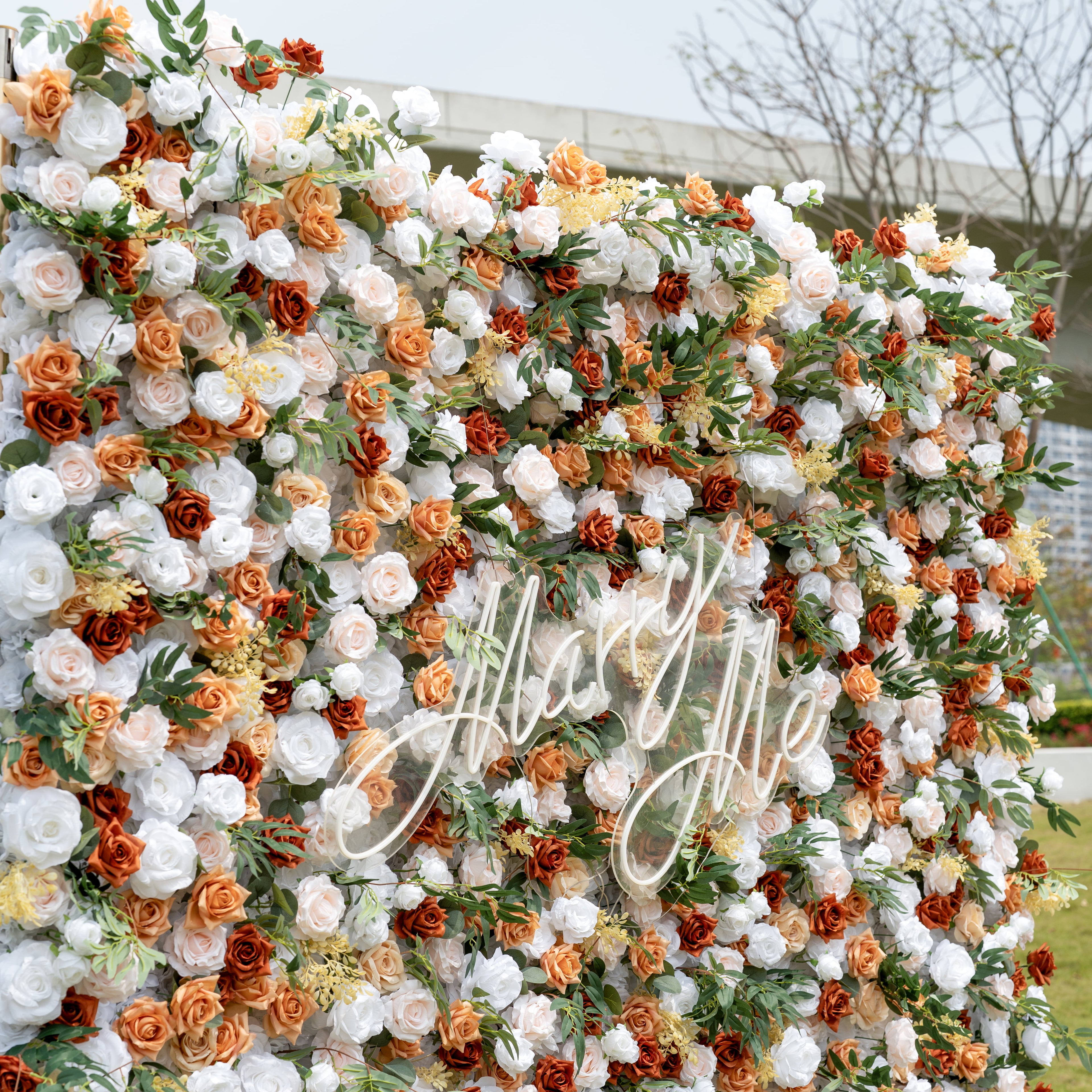 This is a lush, premium artificial flower wall backdrop, brimming with lifelike blooms in a warm terracotta and cream palette. It features burnt orange roses, creamy white peonies, soft blush roses, delicate yellow filler flowers, and vibrant green foliage, centered with an elegant "Marry Me" neon script sign. This dense, realistic, reusable arrangement creates a romantic, showstopping focal point, perfect for wedding proposals, rustic weddings, fall events, and photo booths.