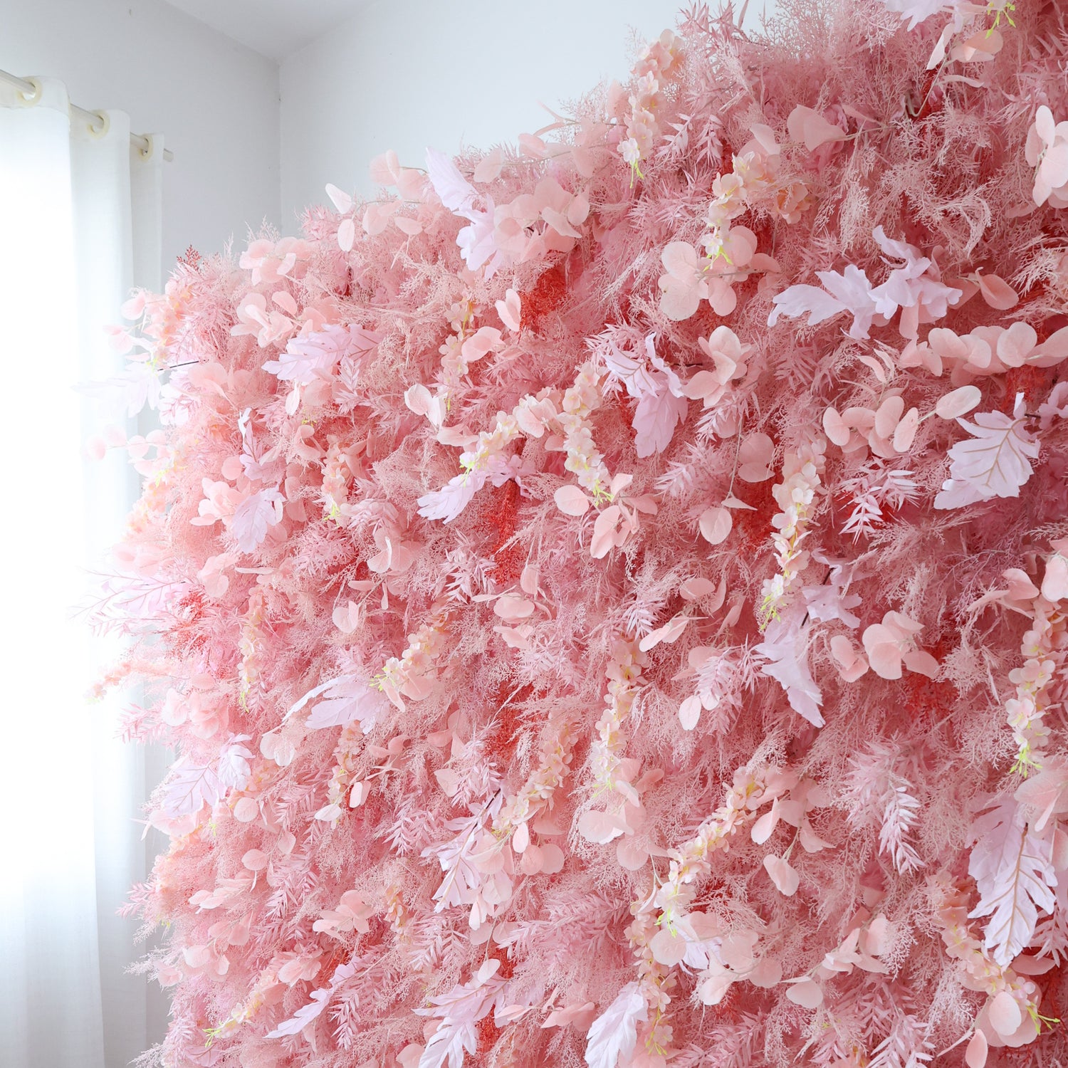 This image presents a large, dense arrangement of artificial pink flowers and foliage indoors. The flowers, in various soft pink shades with delicate petals, are clustered tightly to form a lush, romantic display. Set in a room with white walls and near a window with curtains, the harmonious blend of pink hues and intricate botanical elements creates a gentle, elegant scene, perfect for event decor or as an artistic interior backdrop.