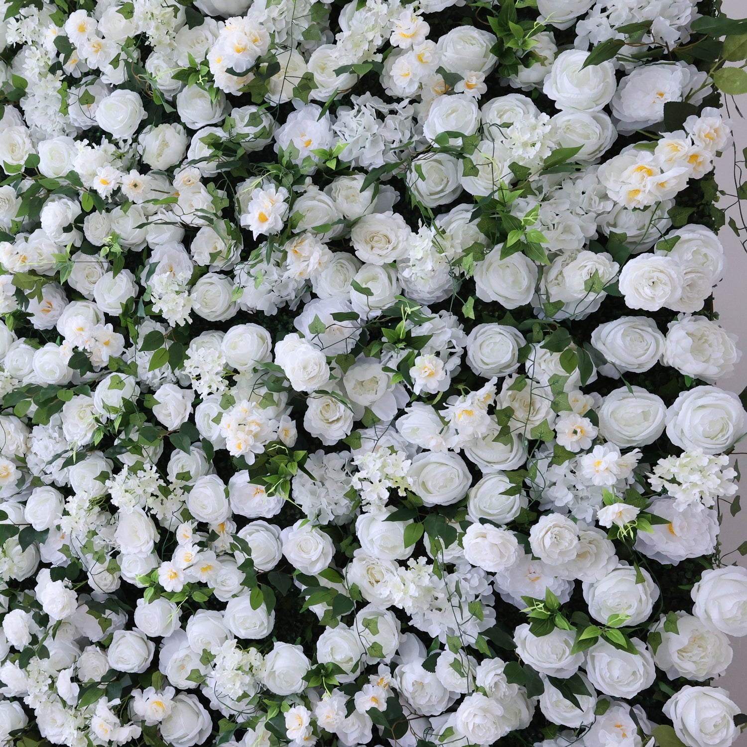 This image showcases a dense, vibrant arrangement of artificial white flowers, mainly roses, interspersed with lush green leaves. The flowers create a rich, elegant tapestry, exuding purity and charm. Perfect for decorative use in events or as an eye-catching interior backdrop, the scene blends pristine white hues and natural-looking foliage to form a visually appealing floral display.