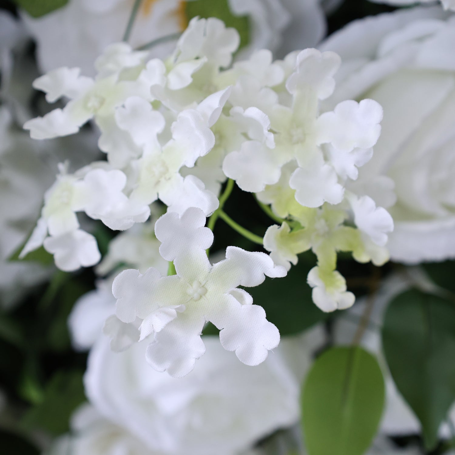 This close - up image showcases a cluster of artificial white flowers with delicate, scalloped petals. The blooms have a soft, almost translucent appearance, and their intricate shape adds an elegant touch. Surrounding the white flowers are hints of green leaves and other white floral elements, creating a harmonious and charming arrangement. The overall scene exudes a sense of purity and grace, perfect for decorative purposes in events or as part of a floral display.