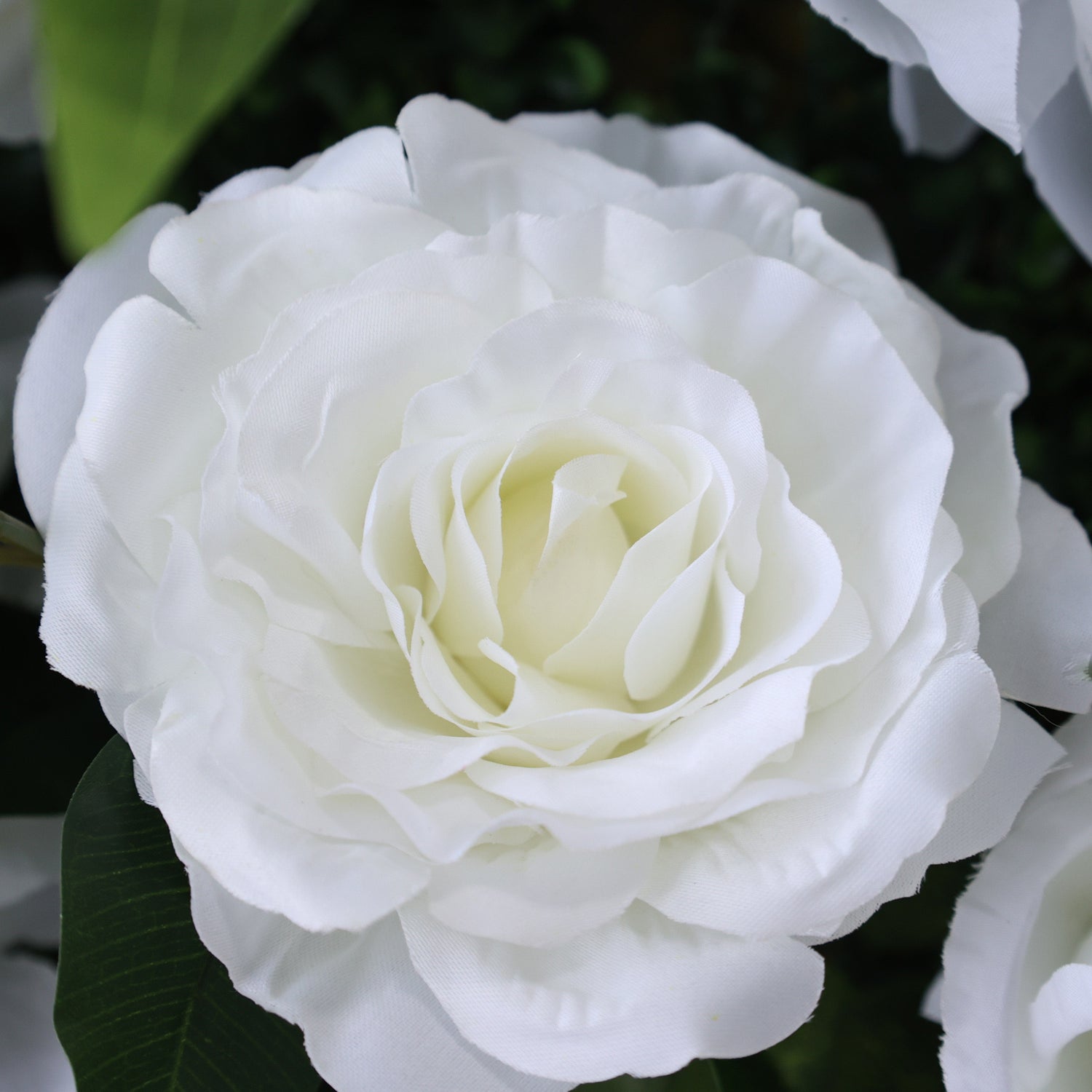 This close - up image showcases a single artificial white rose. The flower features numerous delicate, layered petals that create a lush and full appearance. The petals have a soft, slightly textured surface, mimicking the look of real rose petals. The center of the rose is a creamy white, adding depth to the bloom. Surrounding the rose are hints of green leaves, enhancing the natural and elegant feel of the arrangement. The overall scene exudes a sense of purity and grace, perfect for decorative purposes i