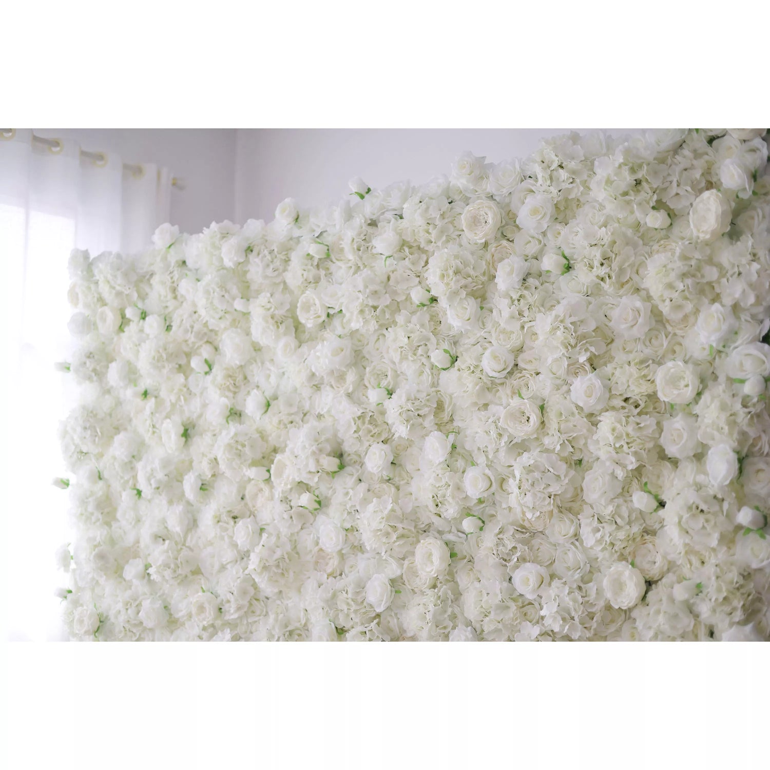 This image presents a dense arrangement of white artificial flowers, creating a lush and voluminous display. The flowers, including roses and other blooms, are tightly packed, giving a sense of purity and elegance. Set indoors with soft light filtering through a curtain, the scene exudes a serene and sophisticated atmosphere, perfect for decorative purposes in events or as a striking interior backdrop, evoking a feeling of natural beauty and refinement.