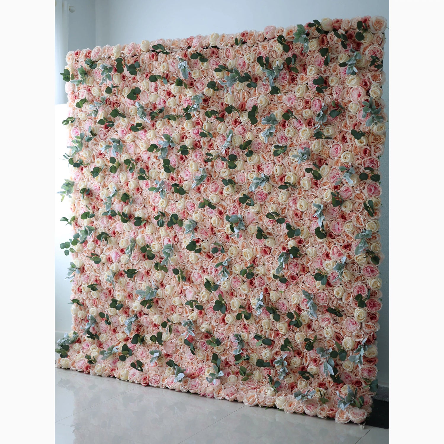 A large artificial flower wall features a base of soft - toned pink roses. Interspersed are green leaves and light - colored foliage, adding texture. The dense, layered arrangement creates a lush, romantic backdrop, ideal for events, blending elegance and natural charm.