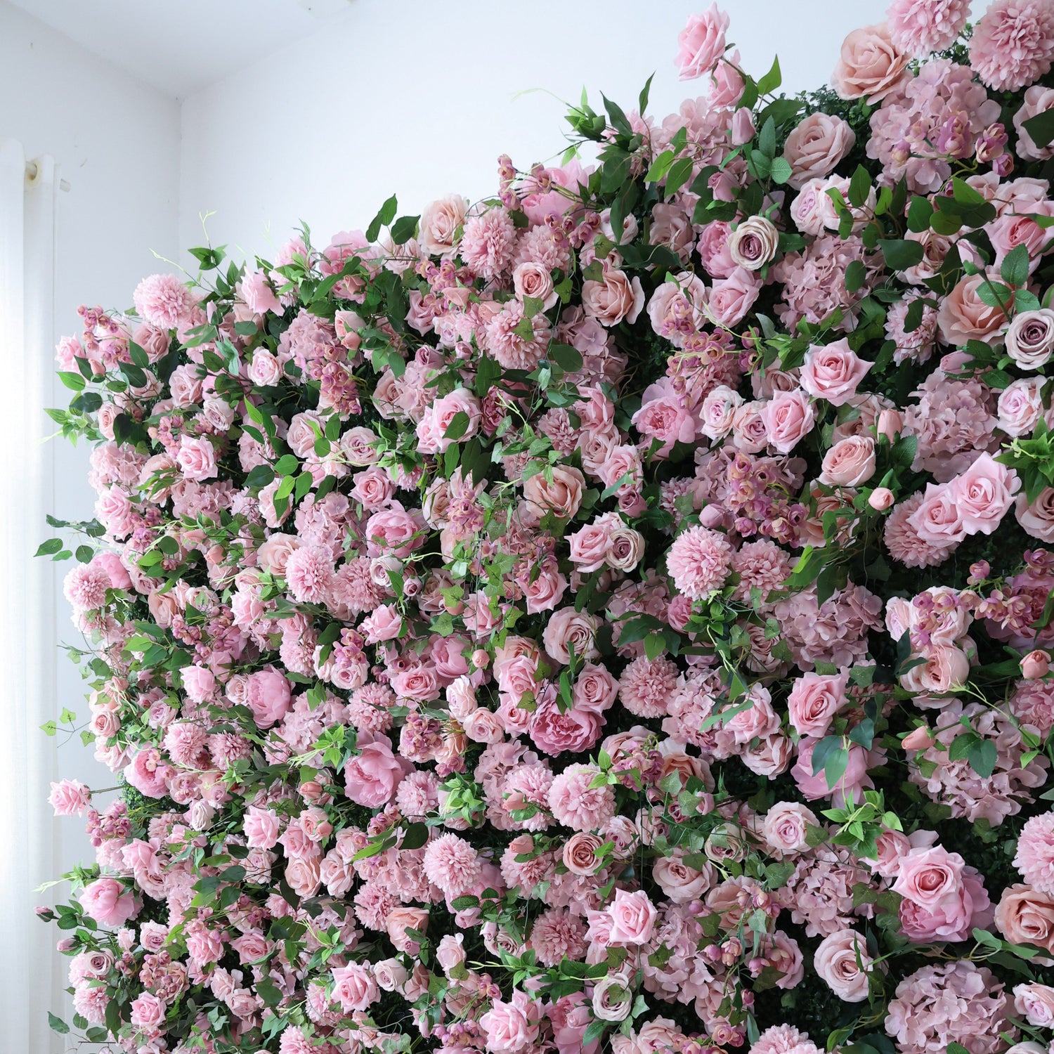 This image showcases a dense, vibrant arrangement of artificial pink flowers, including roses and other blooms, interspersed with lush green leaves. The flowers create a rich, romantic tapestry, exuding elegance and charm. Set indoors against a plain white wall, the scene is perfect for decorative use in events or as an eye - catching interior backdrop, blending soft pink hues and natural - looking foliage to form a visually appealing floral display.