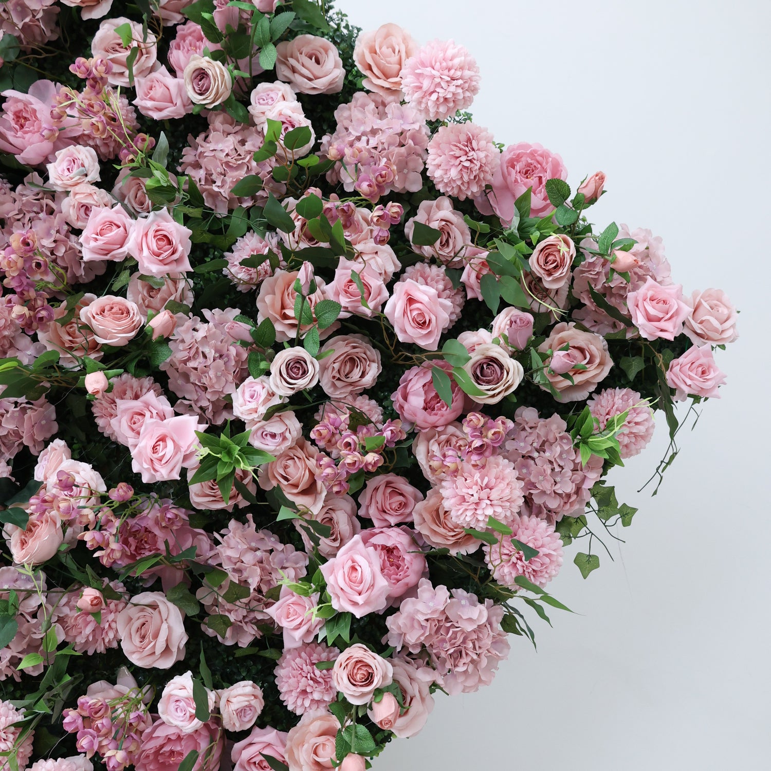 This image displays a dense arrangement of artificial pink flowers, including roses and other blooms, interspersed with lush green leaves. The flowers, in soft pink hues, create a rich, romantic tapestry. Set against a plain white background, the scene exudes elegance and charm, perfect for decorative use in events or as an eye - catching interior floral display.