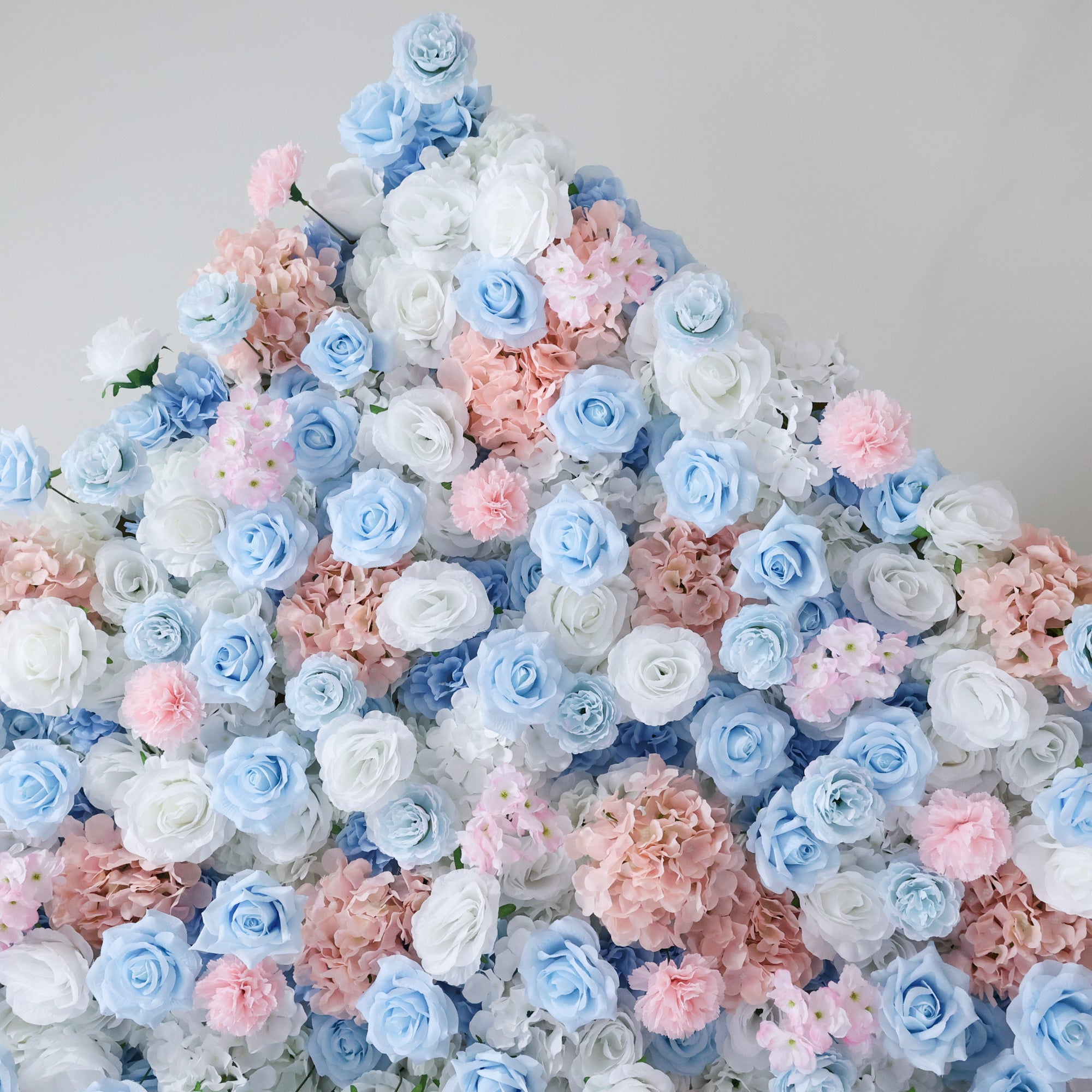 This image showcases a dense arrangement of artificial flowers in soft hues of blue, white, and pink. The blooms, including roses and other floral varieties, are clustered together to form a lush, romantic display. Set against a plain background, the pastel colors and intricate petals create an elegant and charming scene, perfect for decorative purposes in events or as a visually appealing artistic piece.