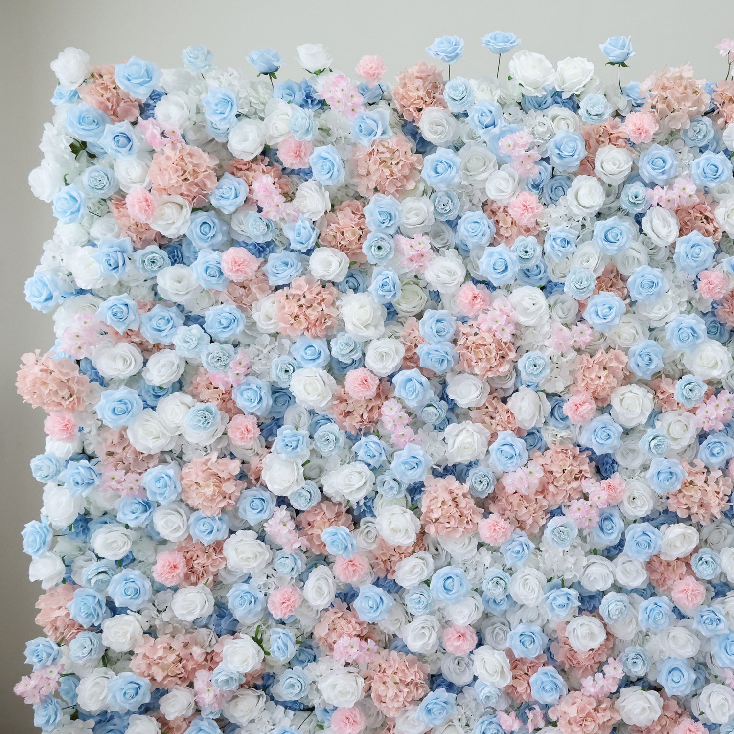 This image showcases a dense arrangement of artificial flowers in soft hues of blue, white, and pink. The blooms, including roses and hydrangeas, are clustered together to form a lush, romantic floral wall. Set against a plain background, the pastel colors and intricate petals create an elegant and charming scene, perfect for decorative purposes in events or as a visually appealing artistic piece.