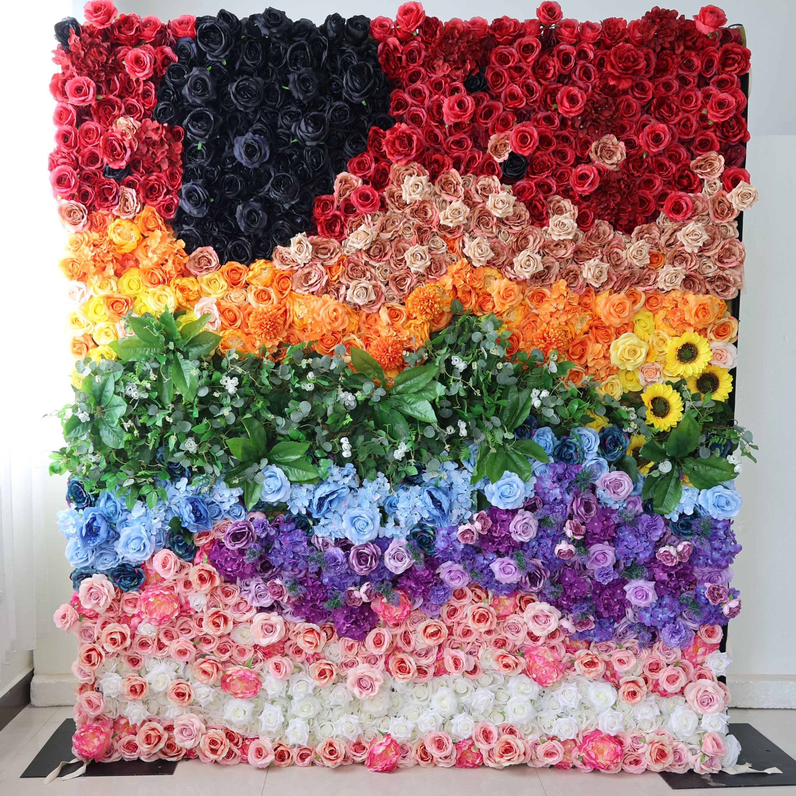 This striking floral wall from Valar Flowers features a bold, inclusive pride flag design crafted entirely from artificial blooms. It showcases distinct sections of deep red, a dramatic black rose accent, warm peach and orange gradients, lush green foliage, cool blue and purple blooms, and soft pink and white roses. The dense, textured arrangement exudes vibrant, joyful energy, making it a powerful focal point for pride events, celebrations, and inclusive gatherings against a minimalist white backdrop.