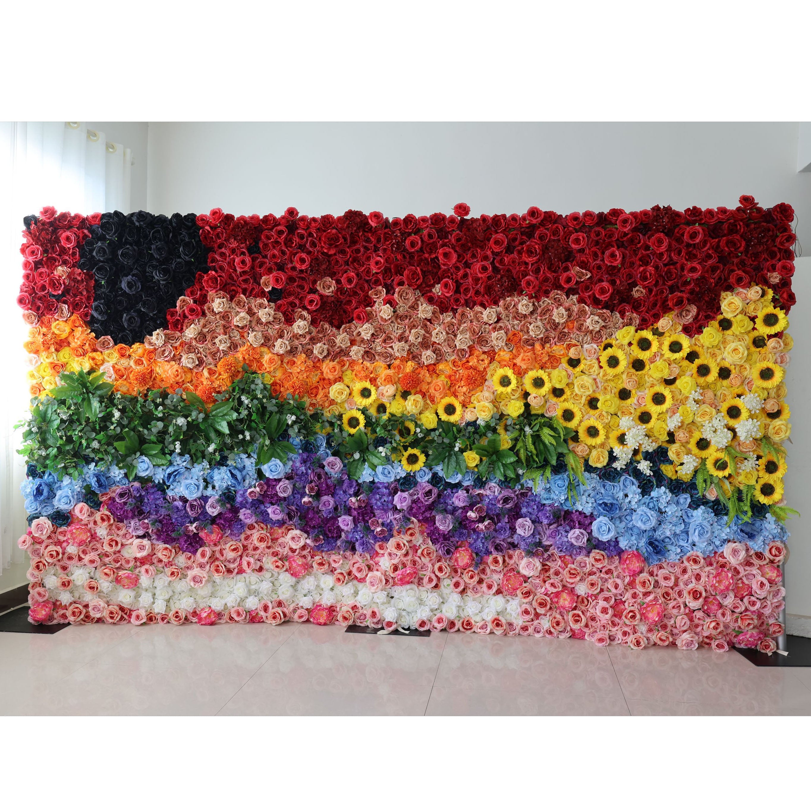 This striking 16x8ft indoor floral wall from Valar Flowers features a bold, inclusive pride flag design crafted entirely from artificial blooms. It showcases a dramatic black rose accent, deep reds, warm peach and orange gradients, sunny yellow sunflowers, lush green foliage, cool blue and purple blooms, and soft pink and white roses. Its dense, textured arrangement exudes vibrant, joyful energy, making it a powerful focal point for pride events, inclusive celebrations, and colorful gatherings against a min