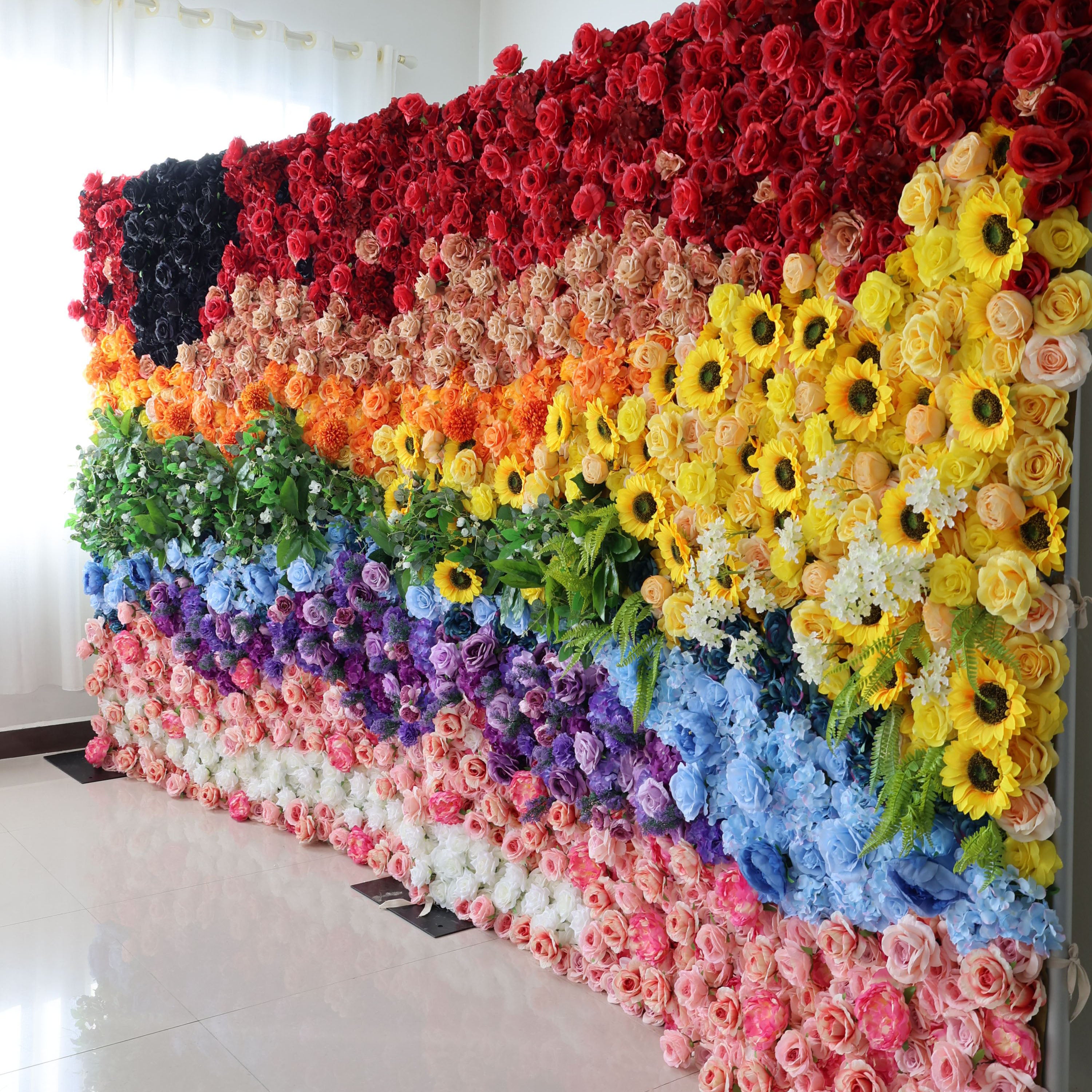 This striking 16x8ft indoor floral wall from Valar Flowers showcases a bold, rainbow-inspired gradient. It features dense sections of deep red, warm peach, sunny yellow sunflowers, lush green foliage, dreamy blue and purple blooms, and soft pink and white roses, with a dramatic black accent on the far left. The textured arrangement of artificial flowers exudes joyful, vibrant energy, making it a lively focal point for pride events, festivals, or colorful celebrations against a minimalist white backdrop.